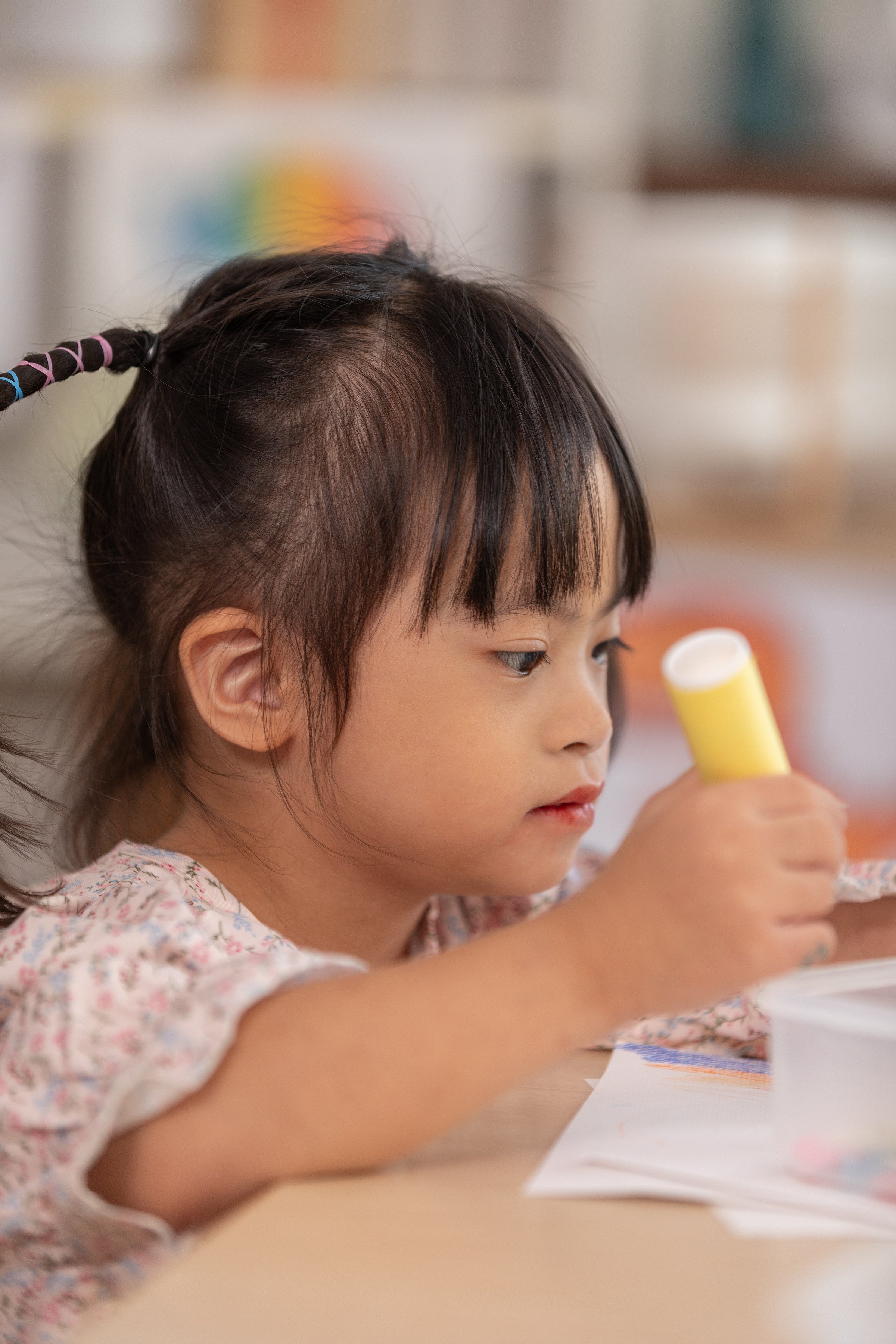 Asian young girl with Down syndrome wearing pigtails, is immersed in arts and crafts project at table. She holds piece of colored paper with focus and concentration, surrounded by crafting materials