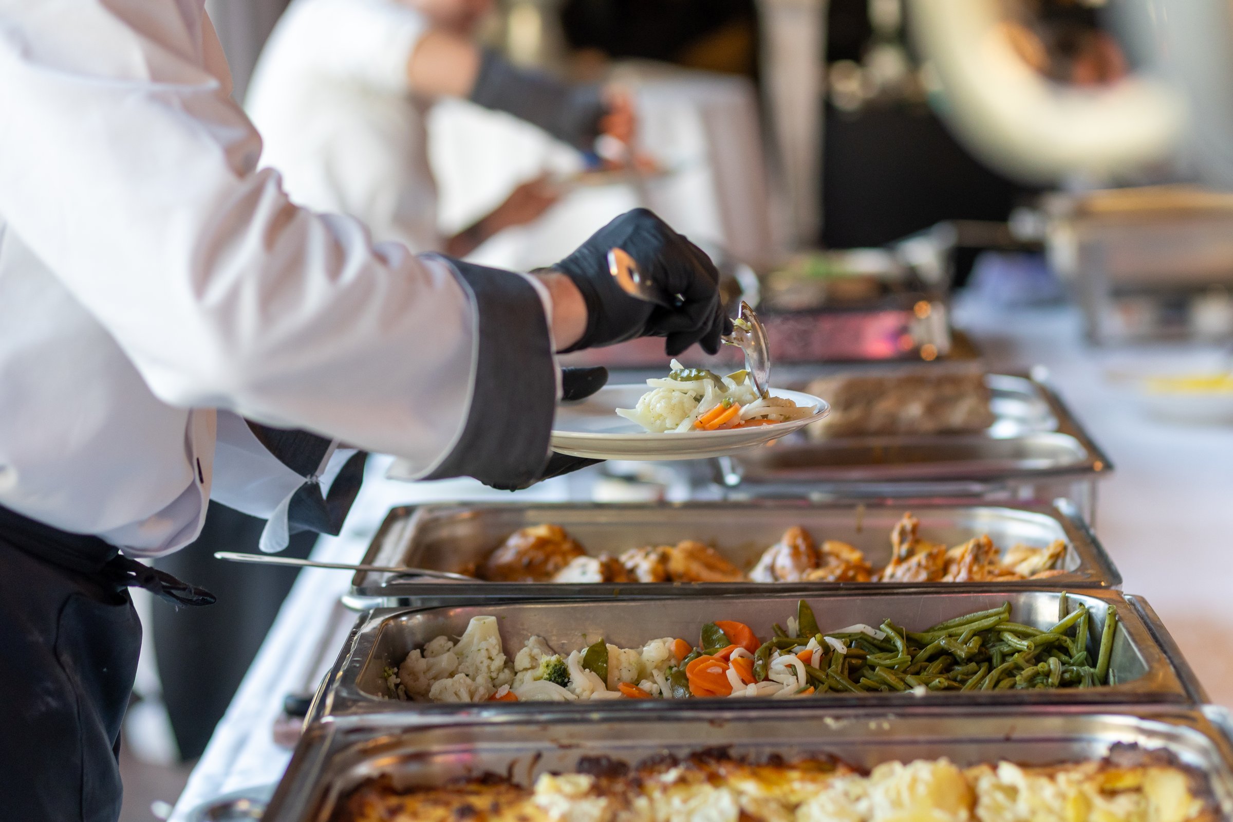 In a spacious and bright event hall, catering staff is actively serving a gourmet buffet. Guests are being offered various dishes, showcasing fresh vegetables and entrees.