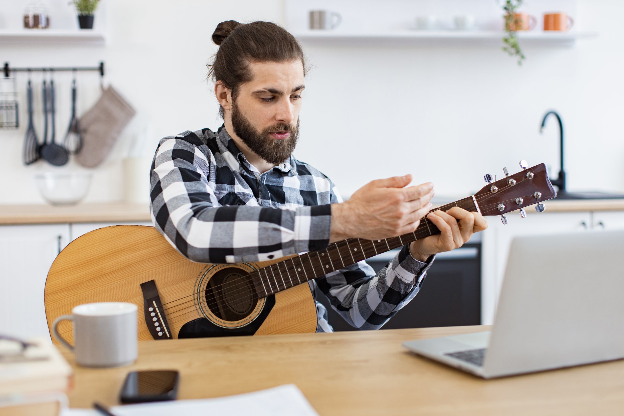 Happy male teaching online guitar lesson using modern technologies. Cheerful Caucasian person holding string instrument while smiling at portable computer webcam in spacious apartment.