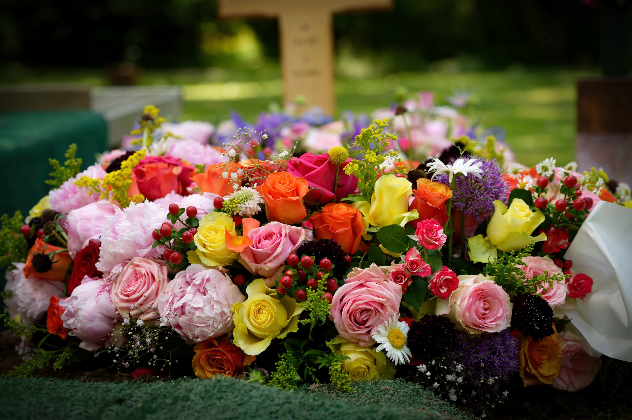 a magnificent floral arrangement of a variety of roses in pink, orange, yellow and red on a grave with a wooden cross in the blurred background
