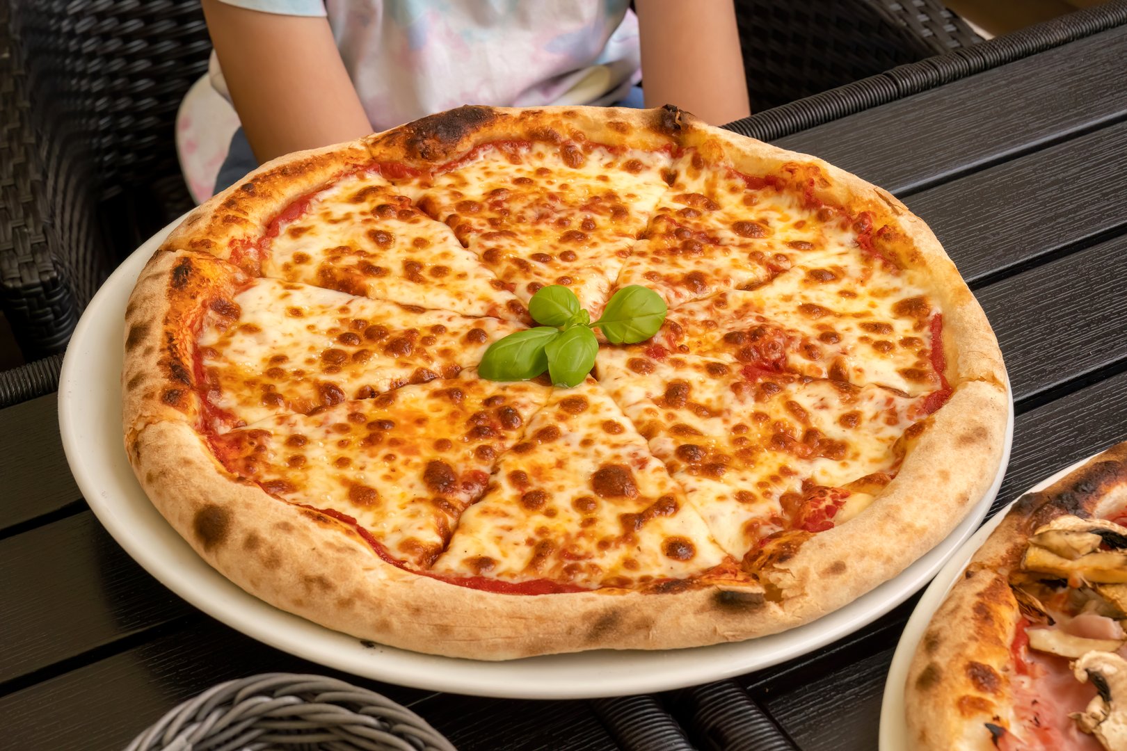 Classic Margherita pizza without additional toppings on the table served in a restaurant, pizzeria, closeup shot thick layer of melted cheese, high angle, one person sitting at the table, basil leaves