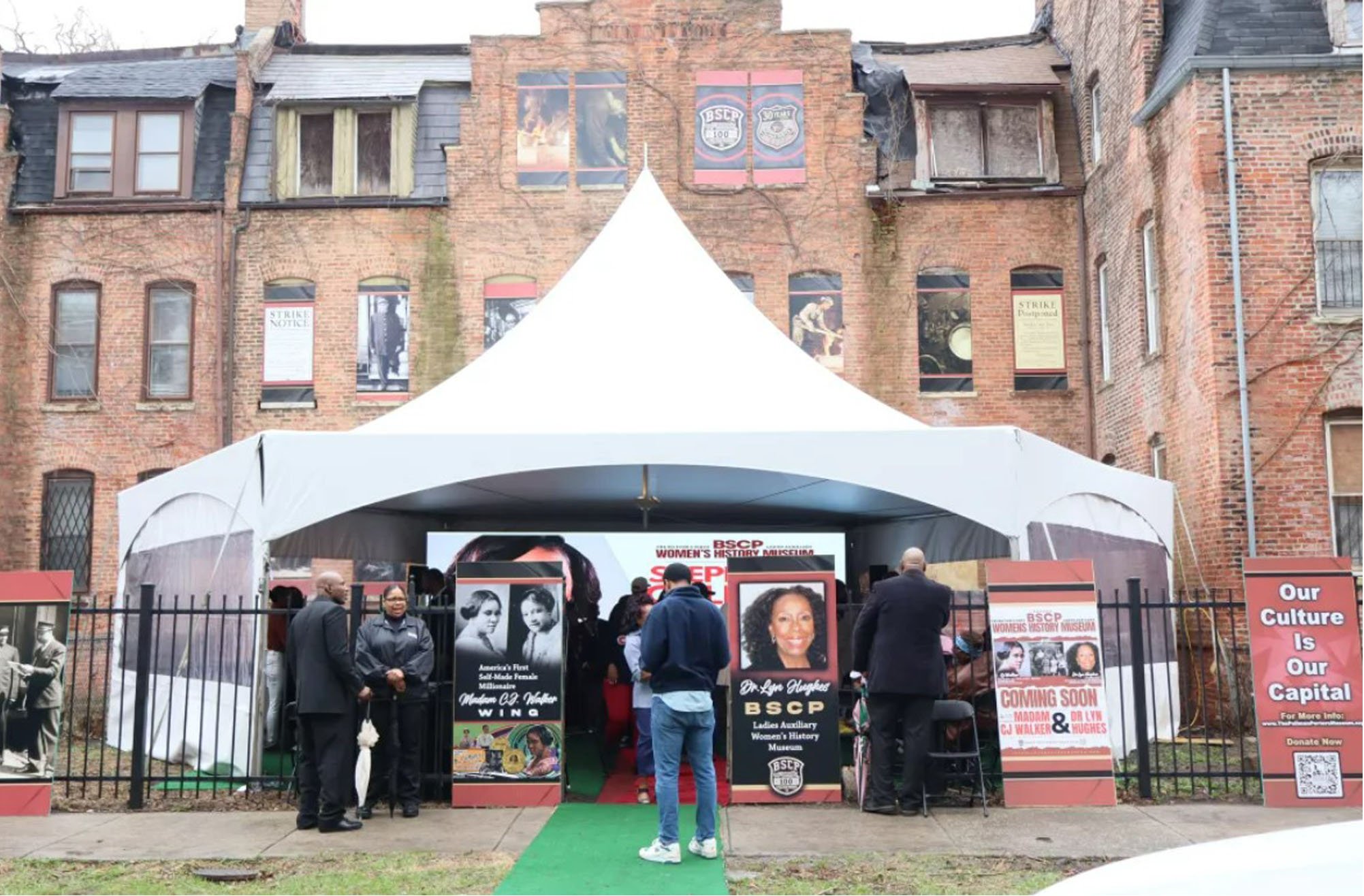 Tent event outside a historic building with a group of people, banners for BSCP, and a sign saying Our Culture is Our Capital.