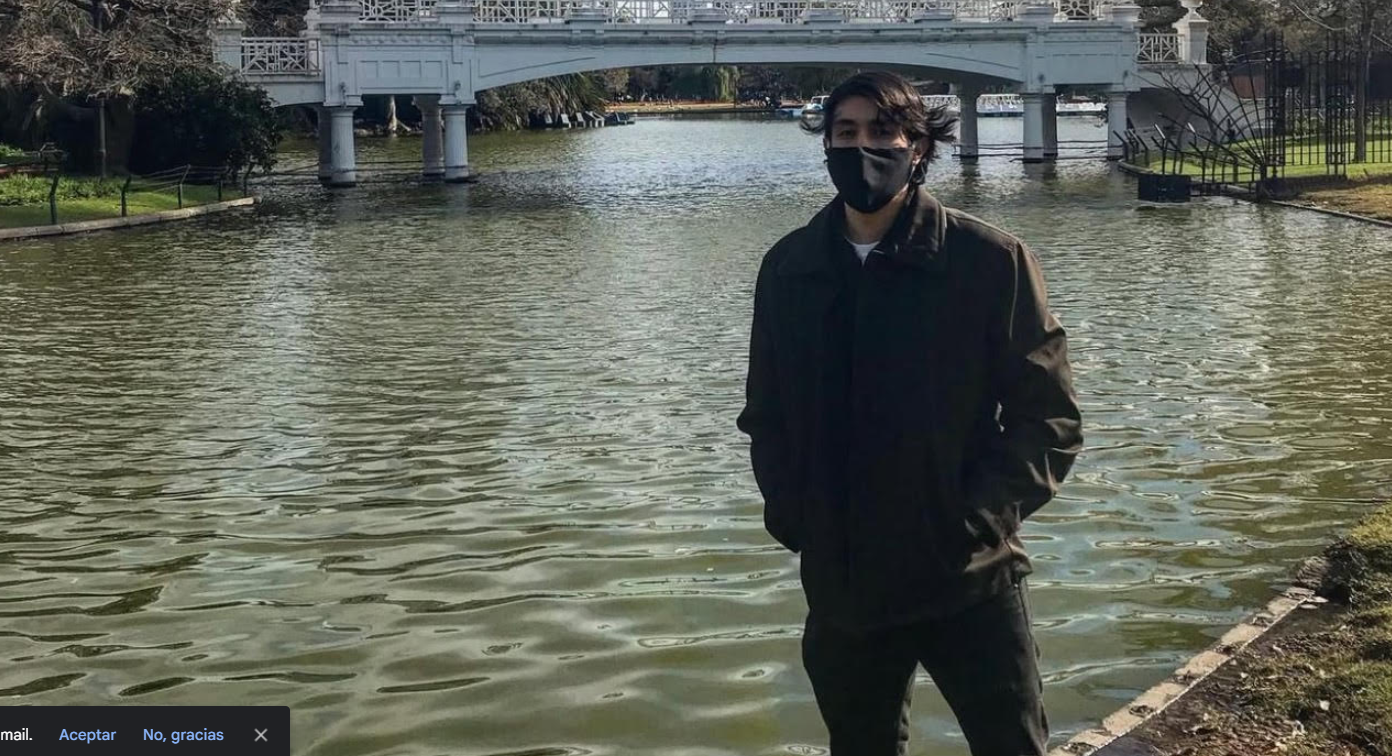 Person wearing a mask standing near a river with a white bridge in the background on a sunny day.