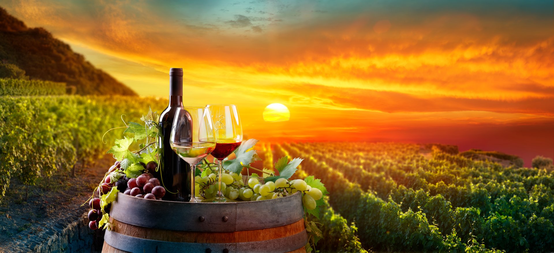 Couple of Wine Glasses With Bottles And Grapes On Barrel In Valley - Landscape At Sunset