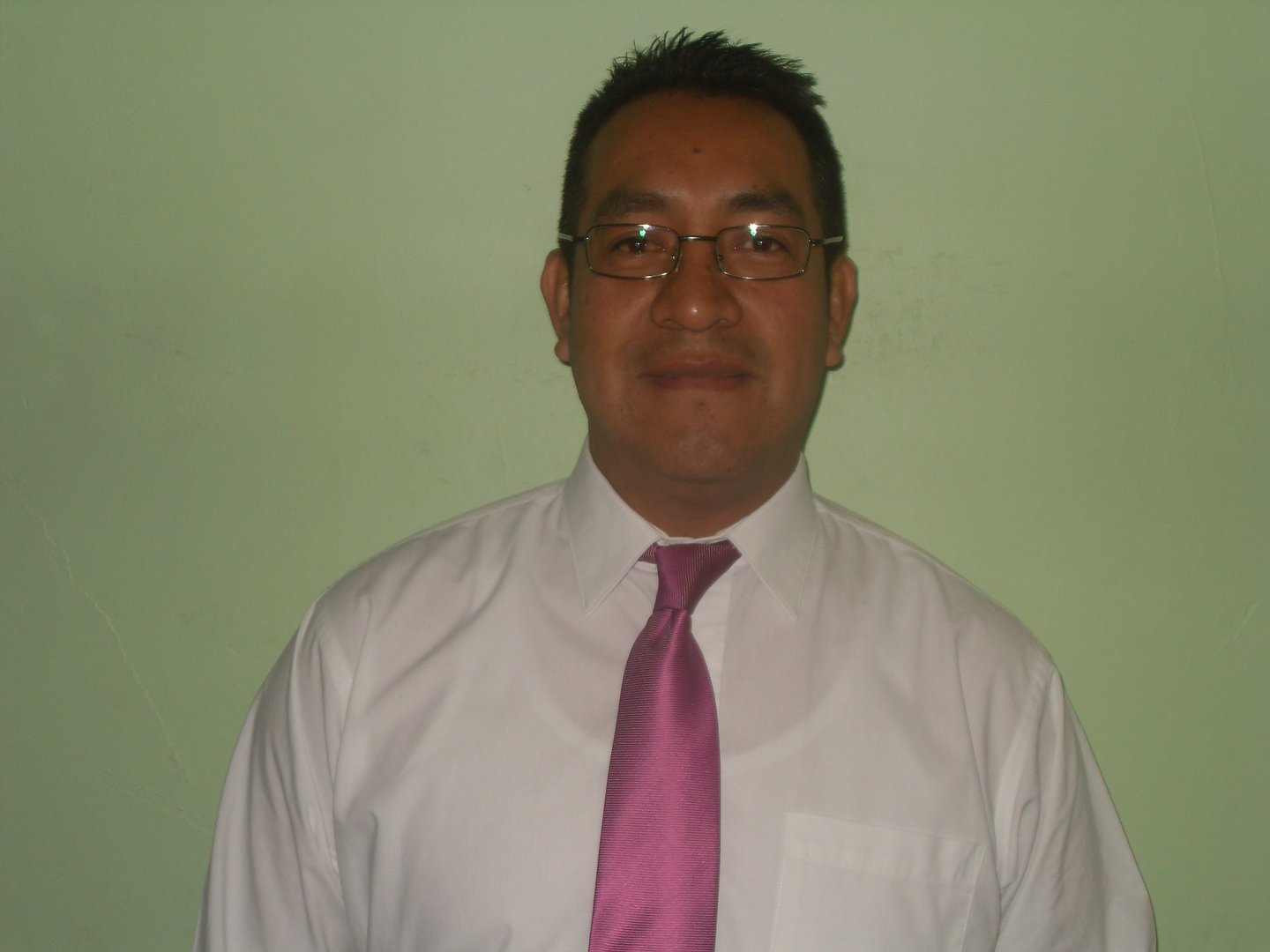 Man wearing glasses, a white shirt, and a pink tie, standing against a green background.