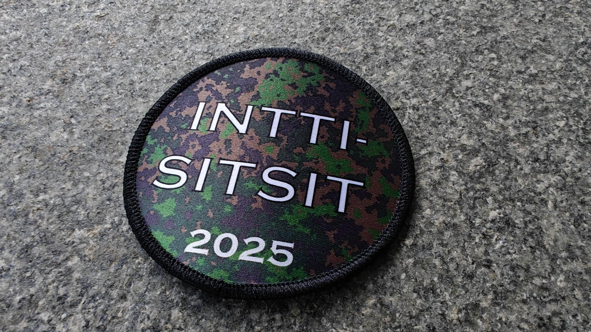 Circular patch with INTTI-SITSIT 2025 on a camouflage background, lying on a textured gray surface.