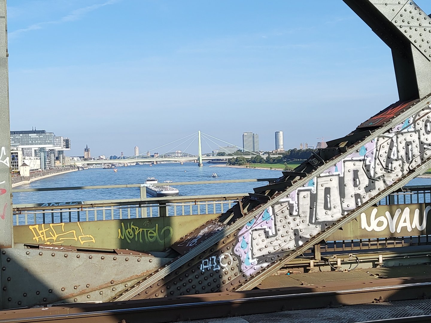 A river view with boats, bridge, and city skyline seen through a graffiti-covered metal structure.