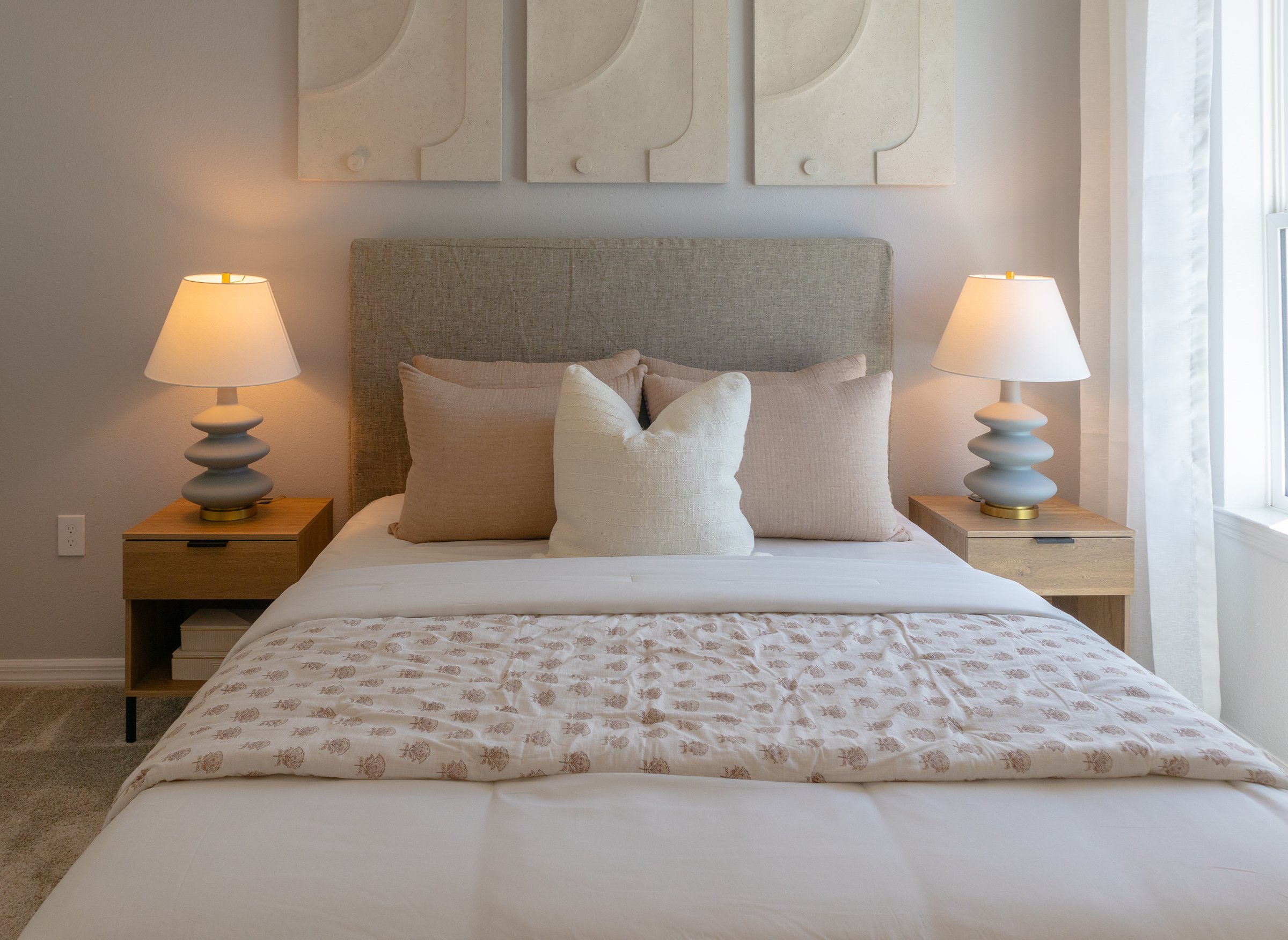 front view of cozy double bed with stacked pillows, blanket, two lamps and night stands with natural light