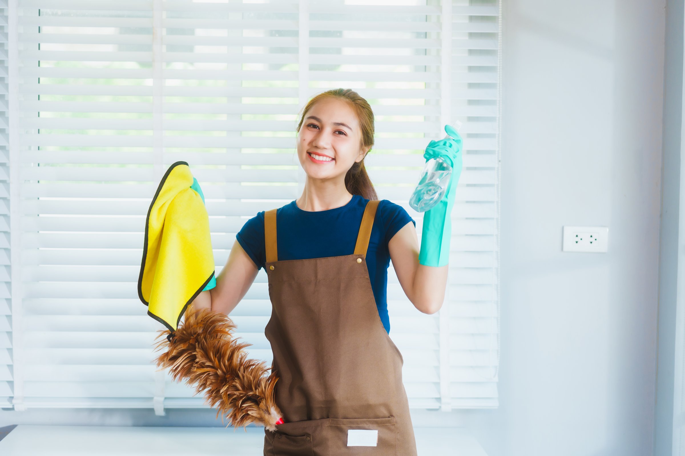 A young Asian housekeeper smiles while dusting window blinds with a feather duster. Wearing gloves and an apron, she creates a clean, fresh, and welcoming home environment in bright natural light.