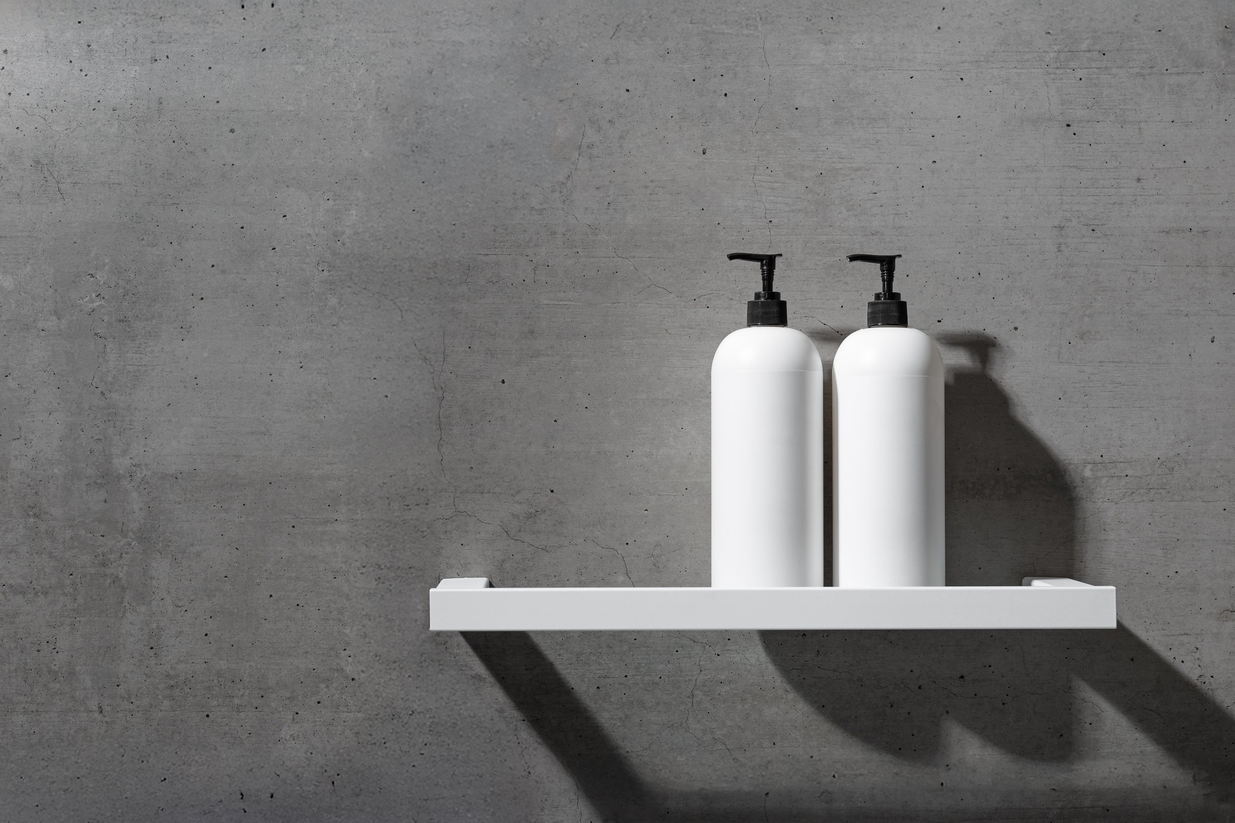 Two minimalist white bottles with black pump caps on white shelf against textured gray concrete background. Neutral tones minimalist interior design bathroom. Skincare, body care, beauty products