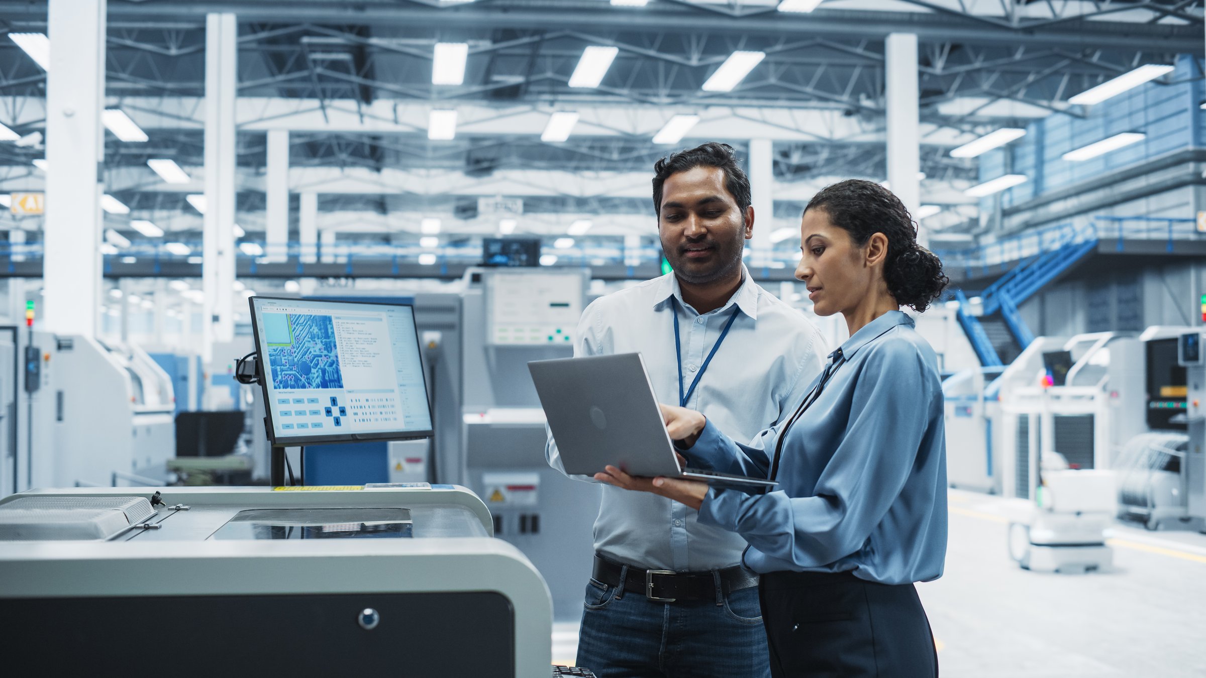 Indian Male Engineer And Hispanic Female Manager Using Laptop Computer And Talking At Electronics Factory. Man Using Soldering Jet Printer, Explaining Process Behind Production Of Circuit Boards