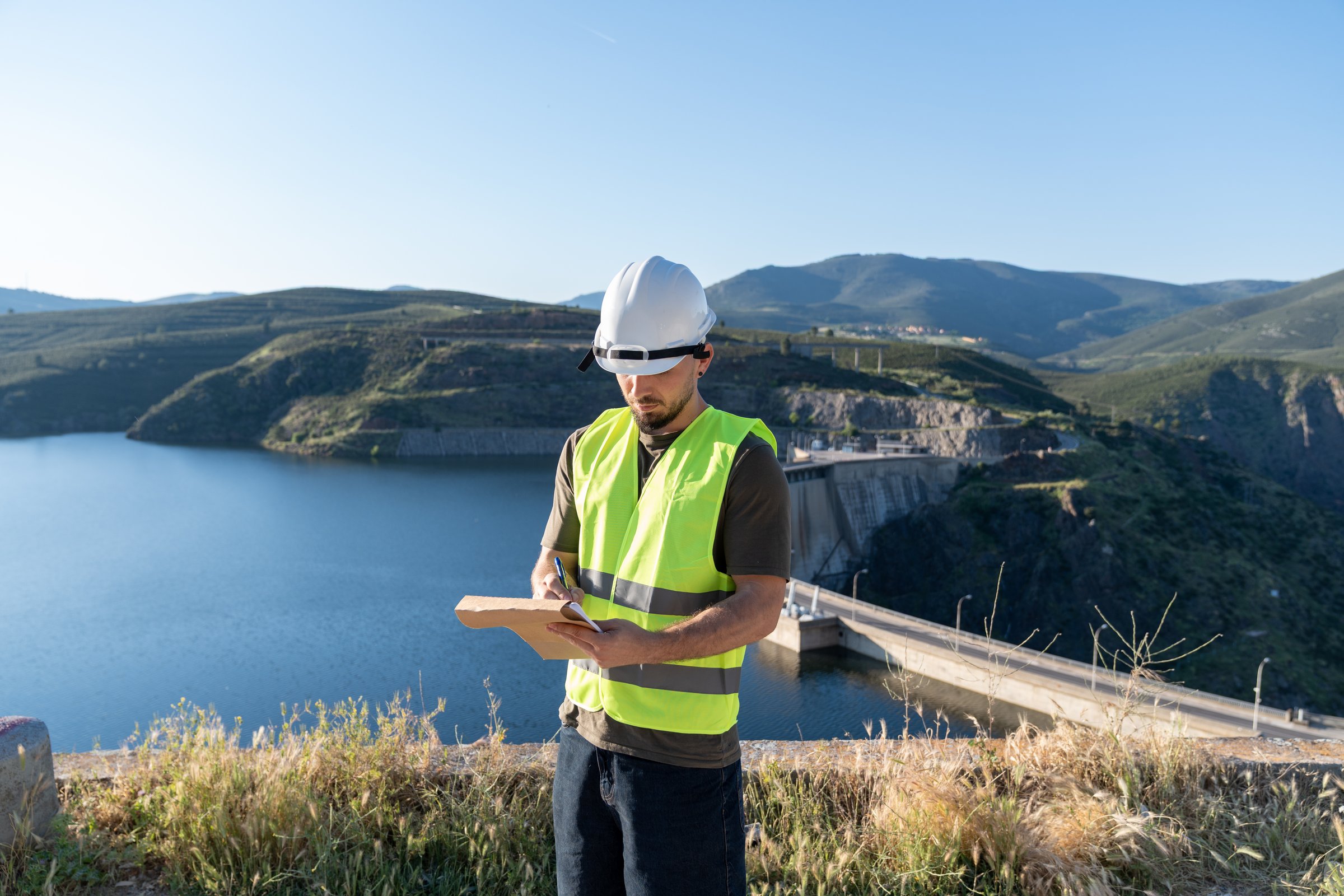 Water technician wearing a safety vest and helmet inspects a reservoir dam and takes notes on a clipboard
