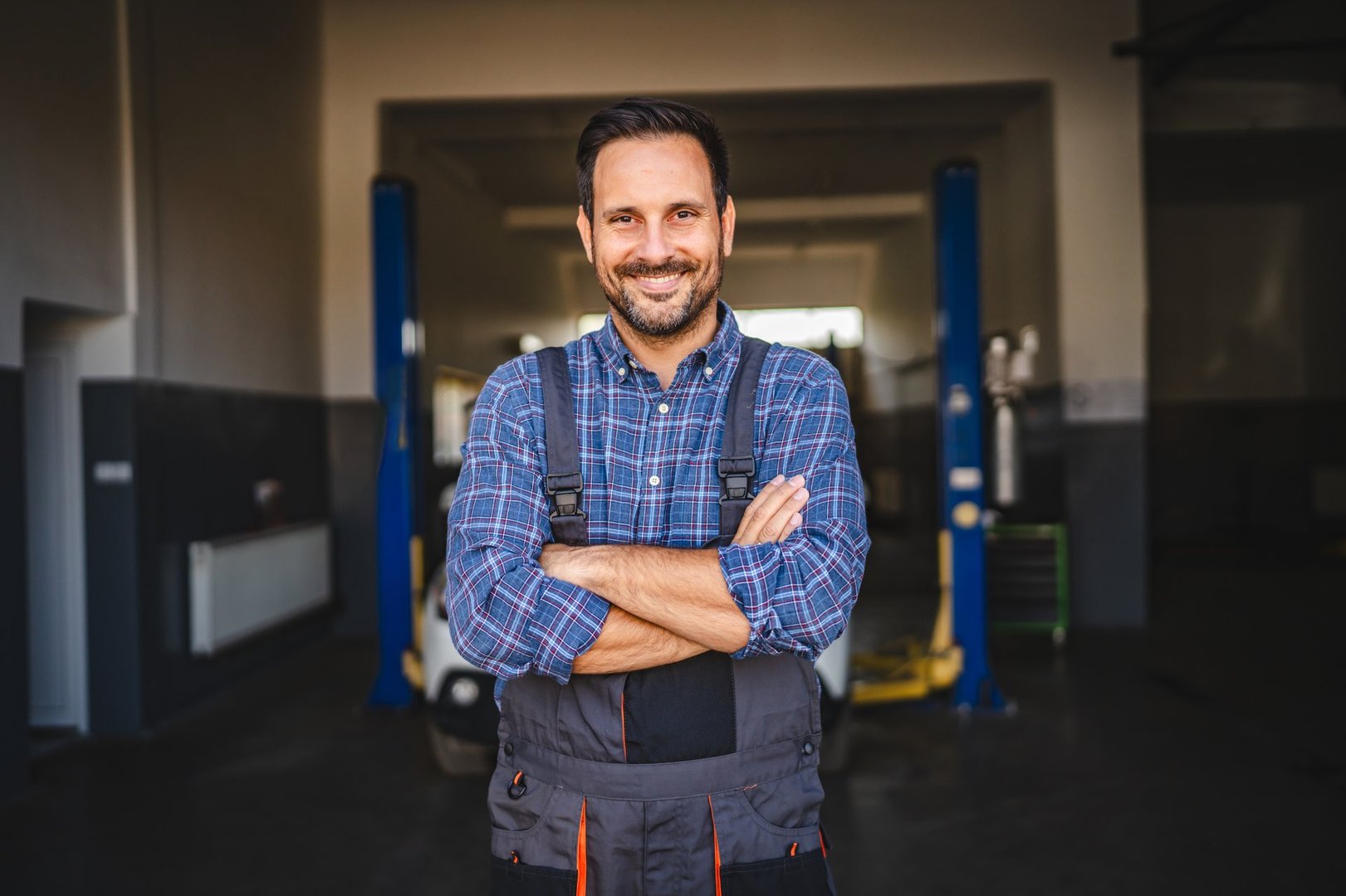 A cheerful mechanic in a plaid shirt stands in front of a garage, arms crossed. Behind him is a car with its hood open, indicating a repair or inspection scene