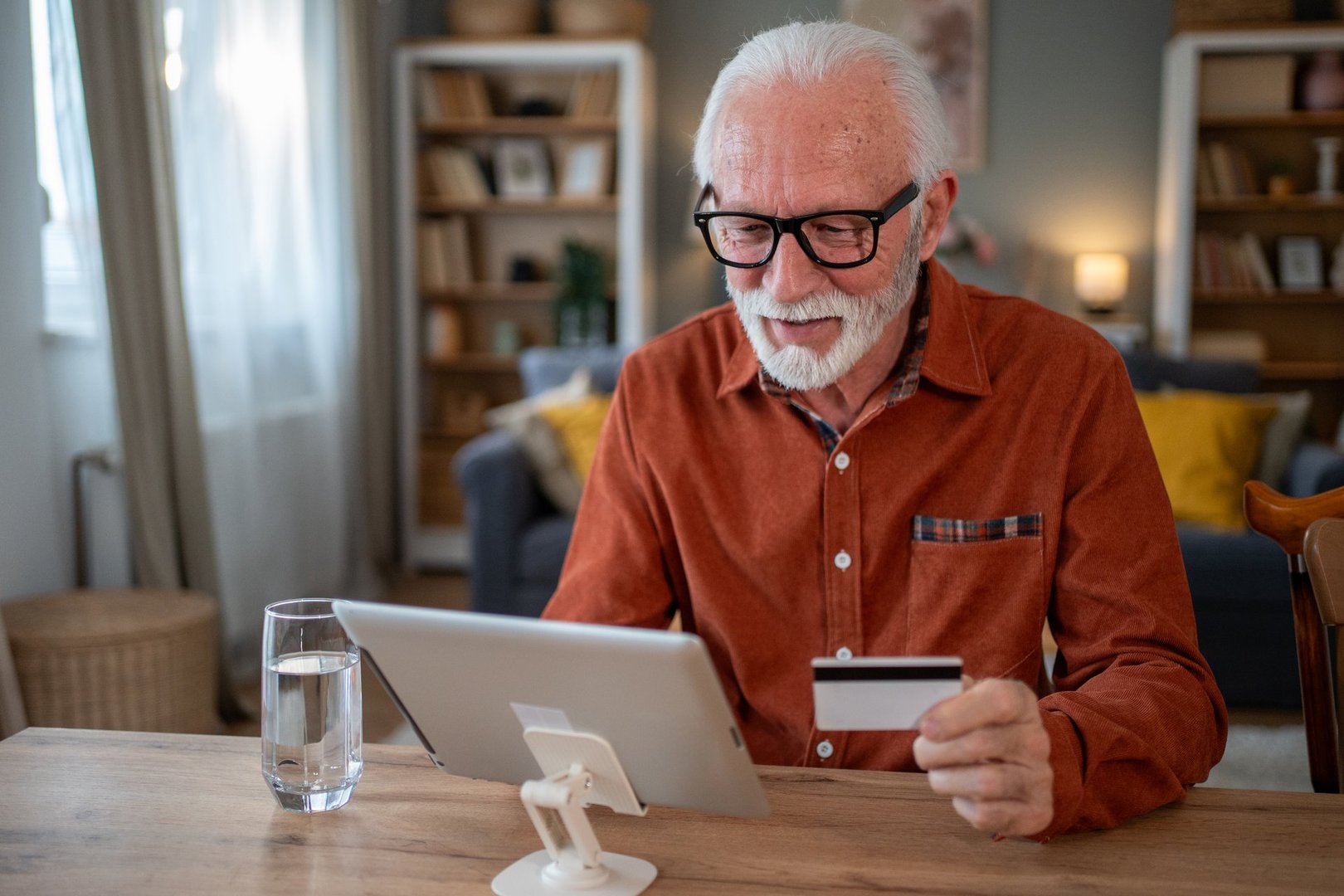 Smiling senior man sitting at a dining table in a cozy home, using a digital tablet and credit card for seamless online shopping, enjoying the convenience of modern technology