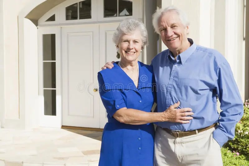 Smiling elderly couple standing together in front of a house, both wearing casual clothing.