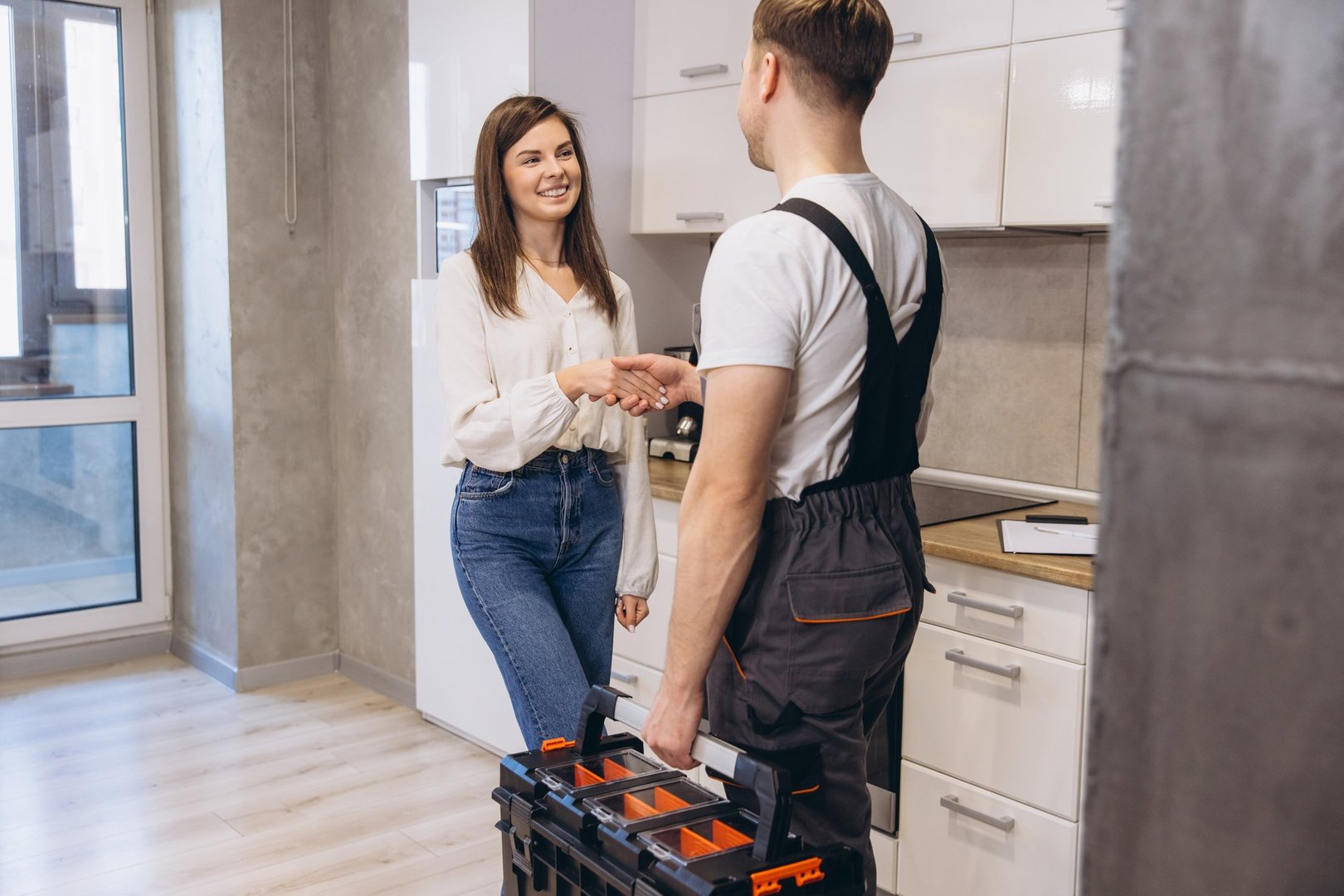 Plumber with toolbox shaking hands with smiling woman in modern kitchen after finishing maintenance work