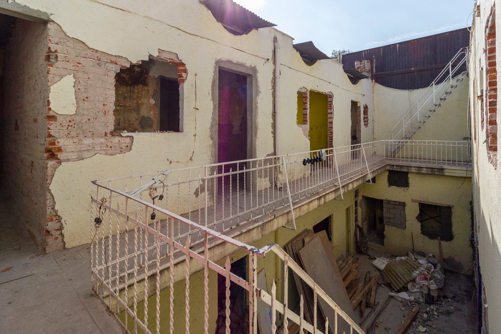 Abandoned building with peeling yellow walls, broken windows, and debris. Metal railings surround a central courtyard.