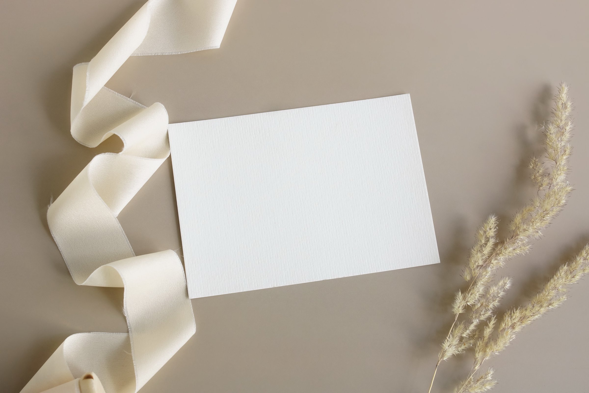 Wedding invitation mockup, empty blank card mock up with white silk ribbon and dry grass top view on beige background. Copy space. Aesthetic minimalist holiday, birthday card template.