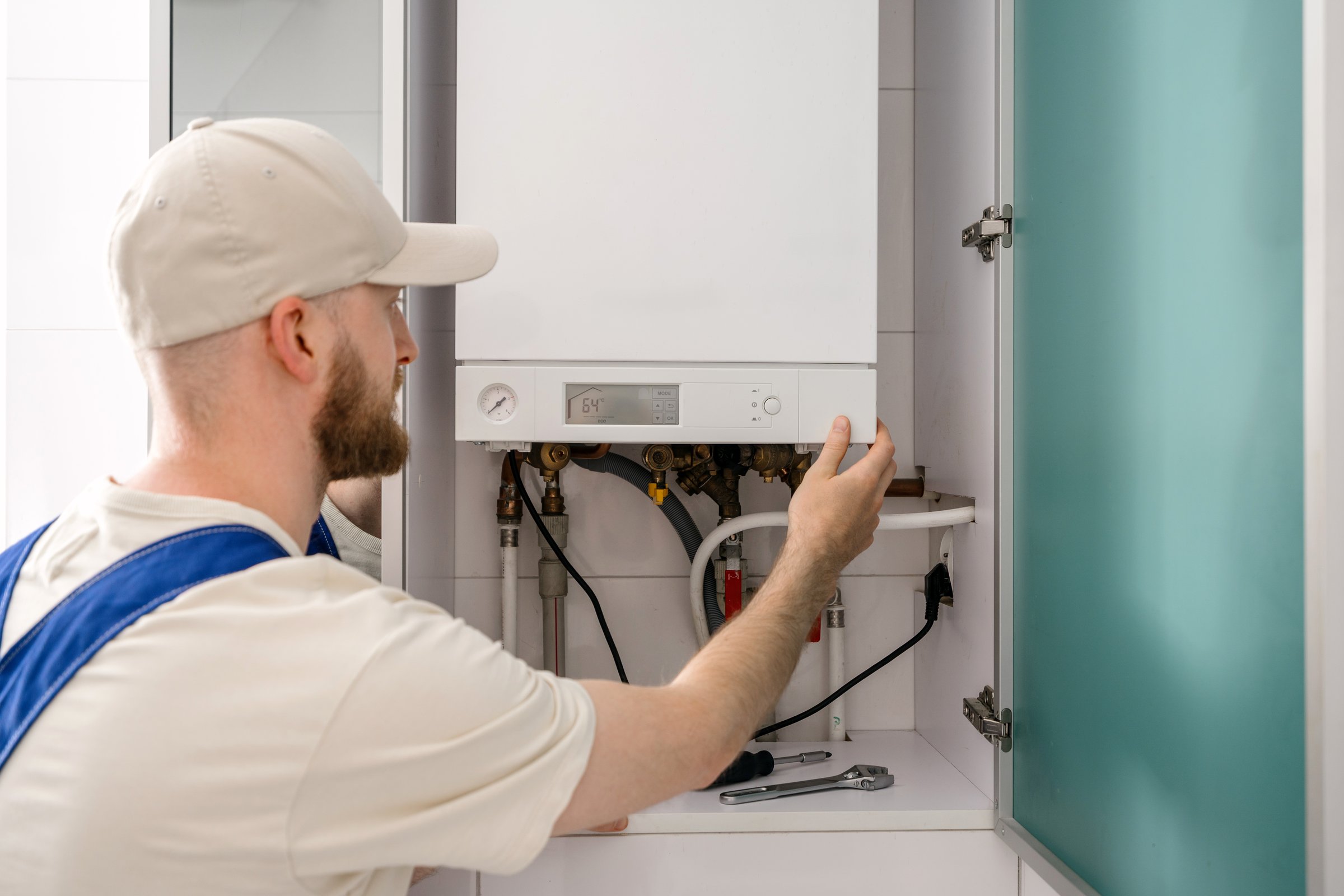 Back rear view of a repairman is inspecting a gas boiler in a bathroom, focusing on maintenance tasks to ensure optimal performance. The worker uses tools to adjust settings and check connections.