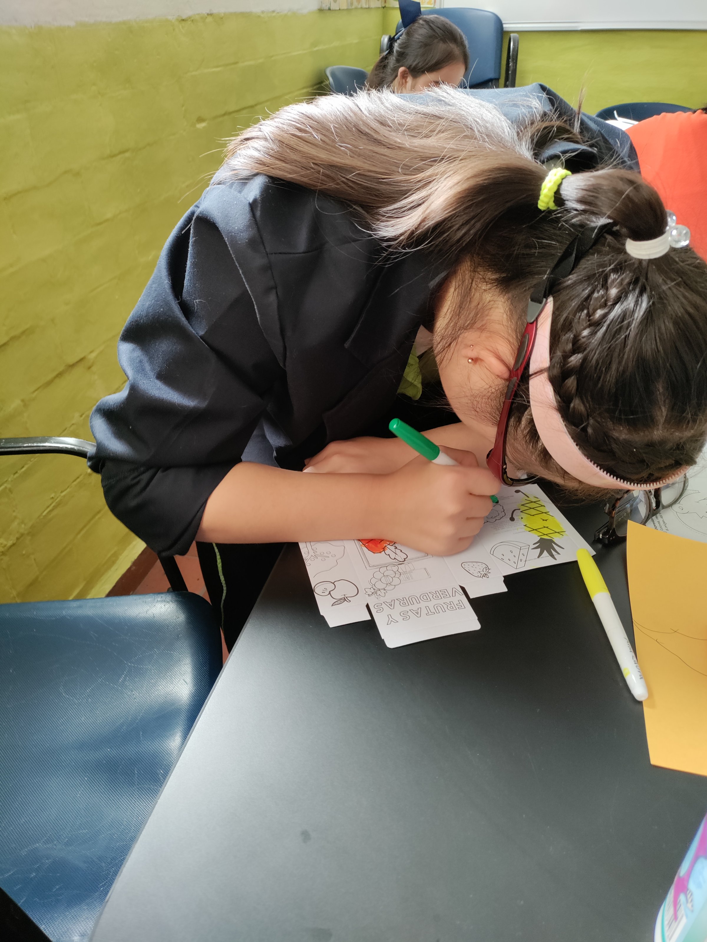 Child coloring a drawing with markers at a table, hair tied back with a headband, in a room with yellow walls.