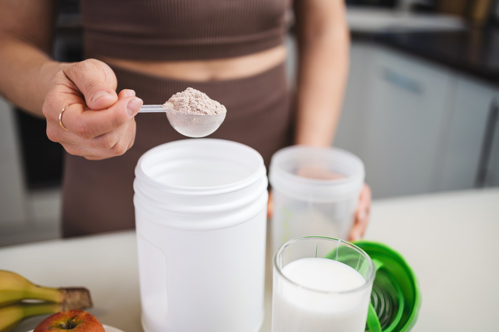 Athletic woman in sportswear preparing protein drink in a kitchen, putting a portion of whey protein powder from a measuring spoon into a shaker in her hand.