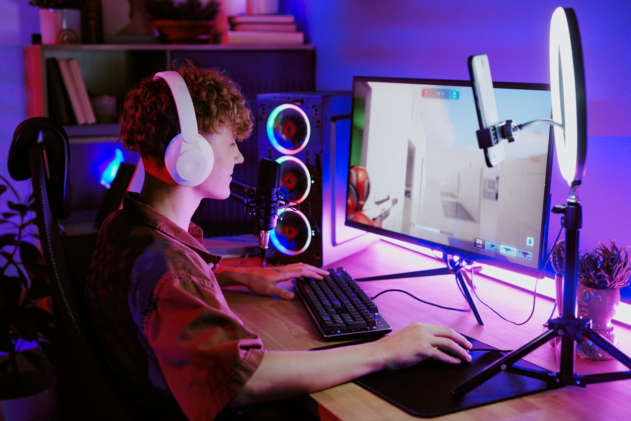 Caucasian teenage boy wearing headphones sitting at desk live streaming video games using desktop computer with microphone and ring light
