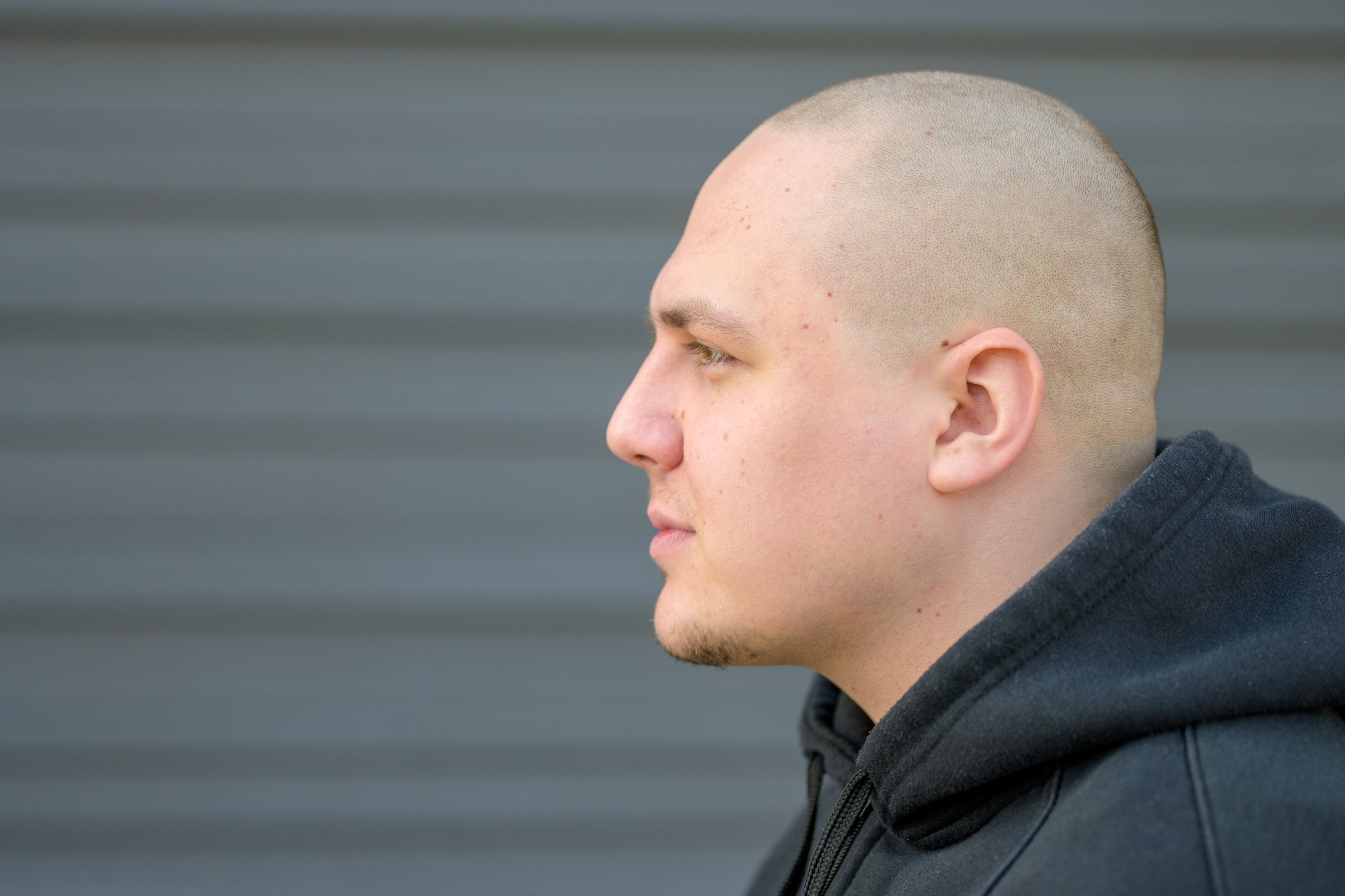 Profile portrait of a modern young man with shaved head looking straight ahead with a deadpan serious expression against a grey wall with copyspace
