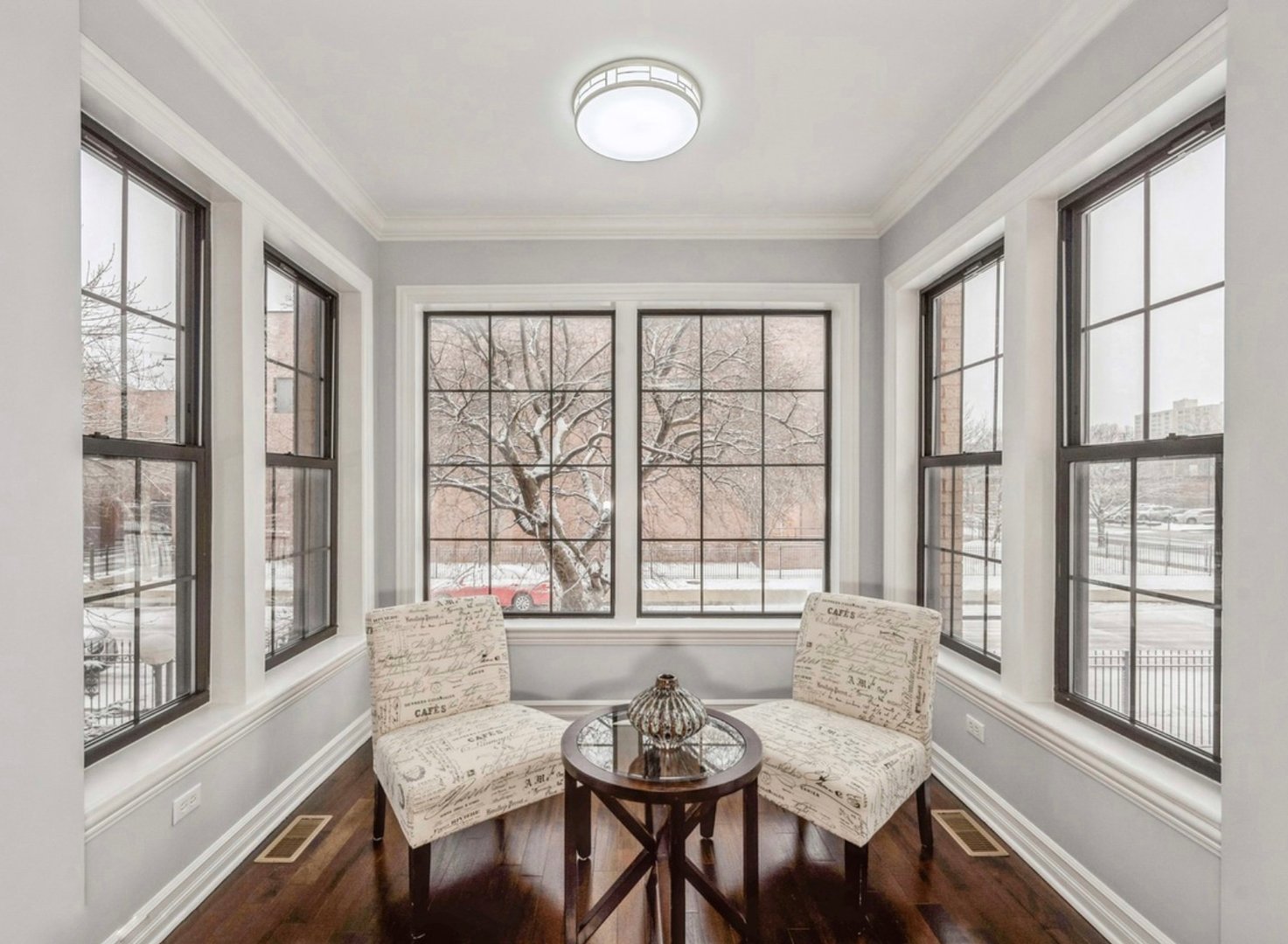 Two upholstered chairs and a round glass table in a bright room with large windows and a snowy view outside.