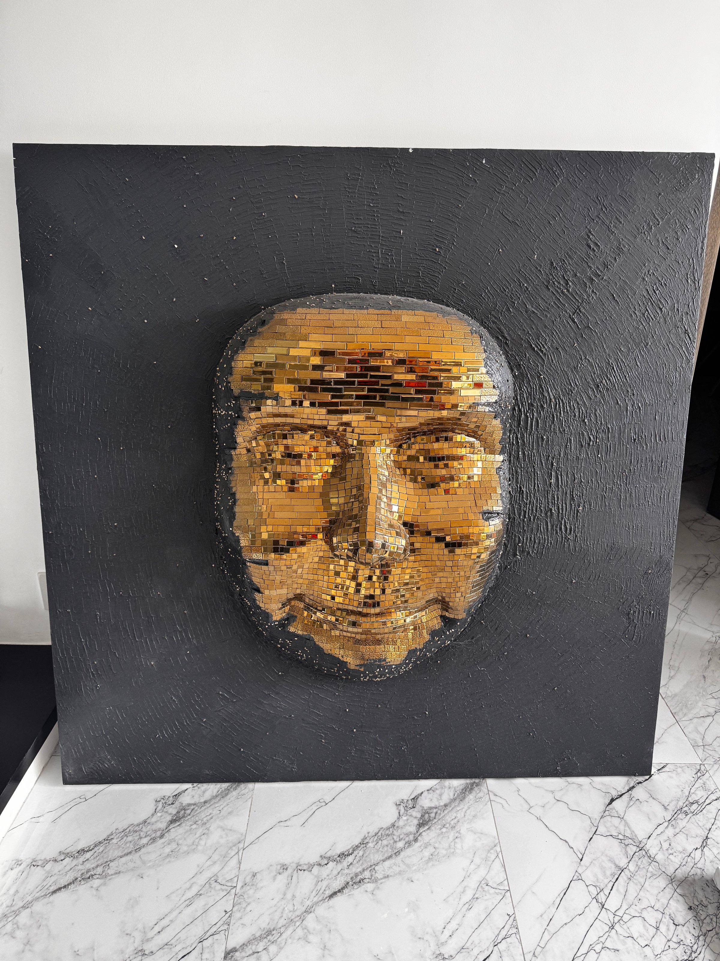 A mosaic art piece of a human face made from golden and black tiles, mounted on a dark textured background.