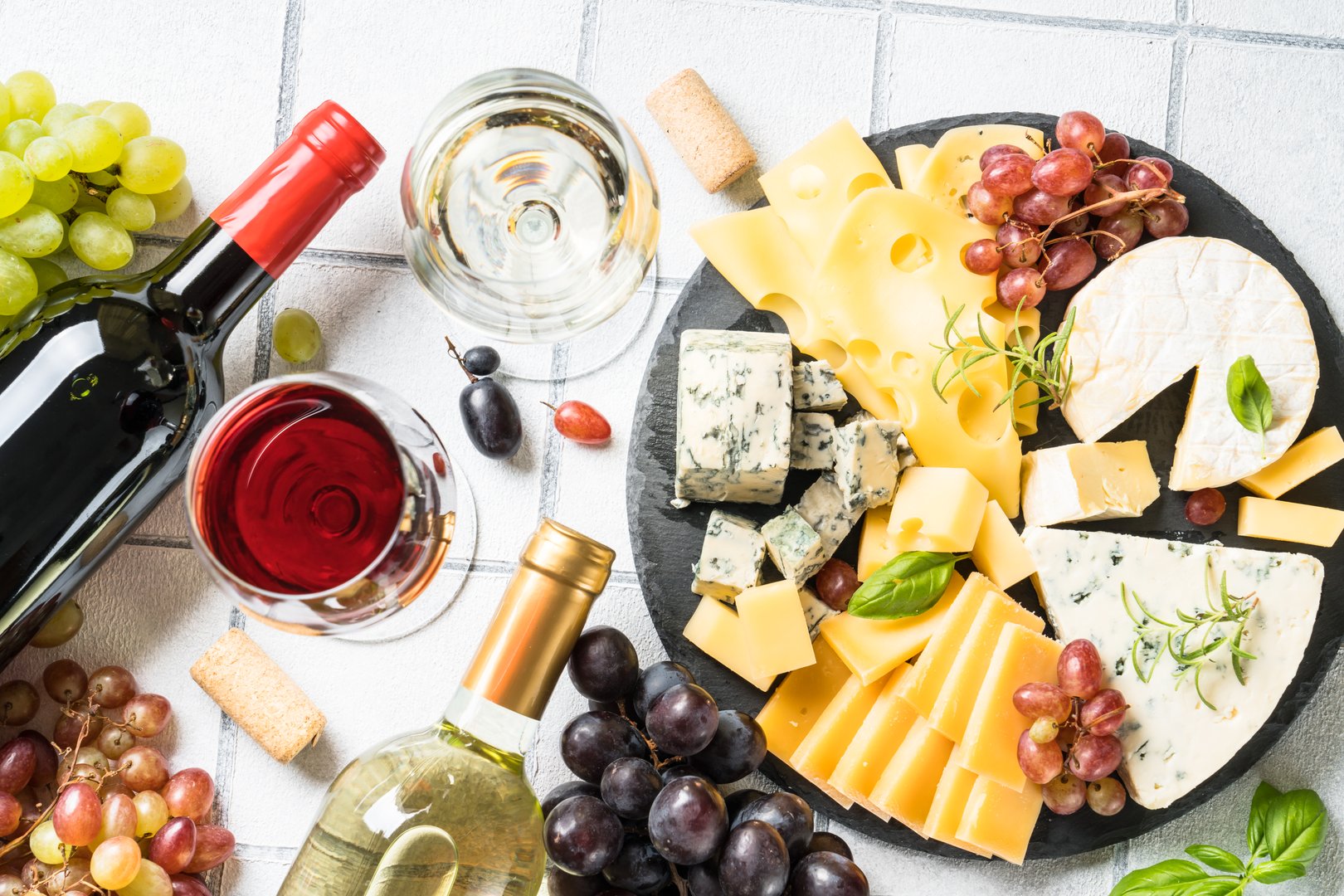 Cheese platter with craft cheese assortment and wine glasses at white tile background. Top view.