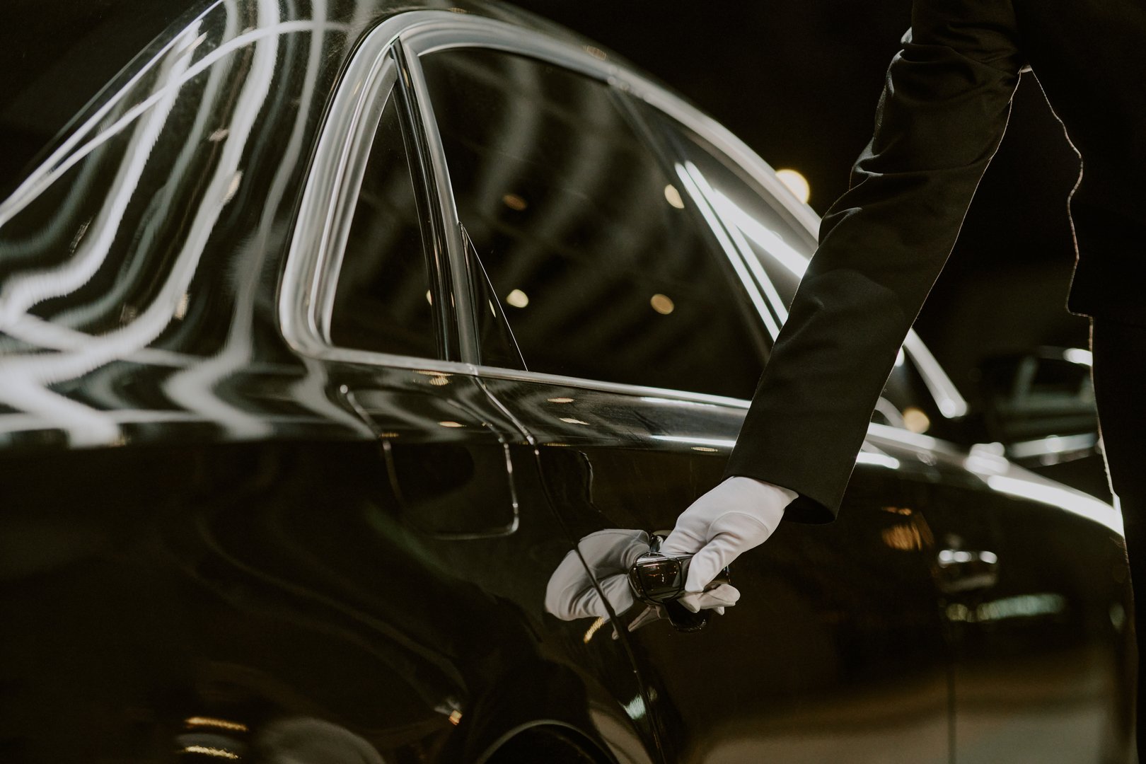 Caucasian middle aged man wearing glove opening car door, only arm and hand visible, luxury vehicle reflecting lights in background, professional chauffeur service concept