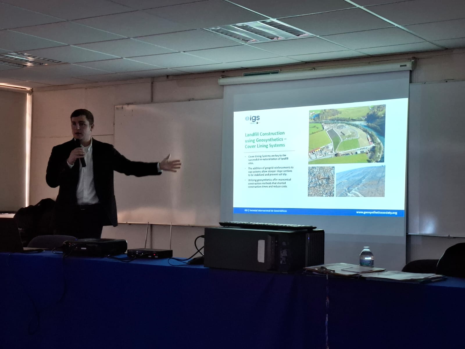 A presenter speaks at a conference, pointing to a projected slide titled 'Landfill Construction Using Geosynthetics.'