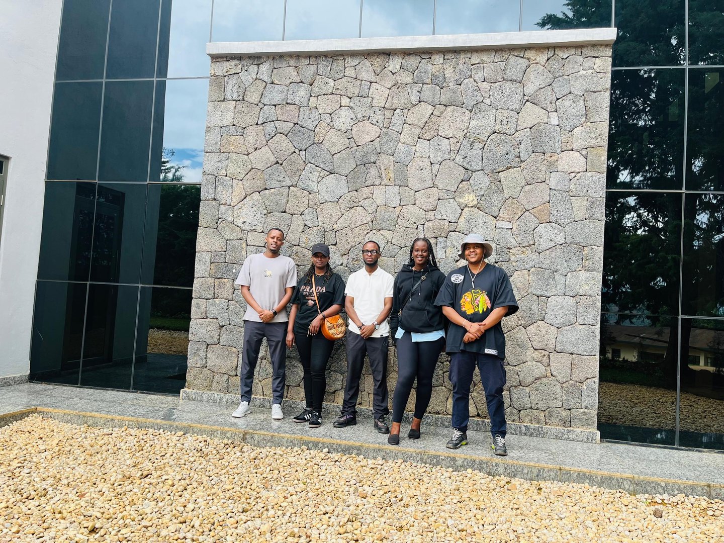 Five people stand on a gravel path in front of a stone wall, posing together, with a partially visible building nearby.