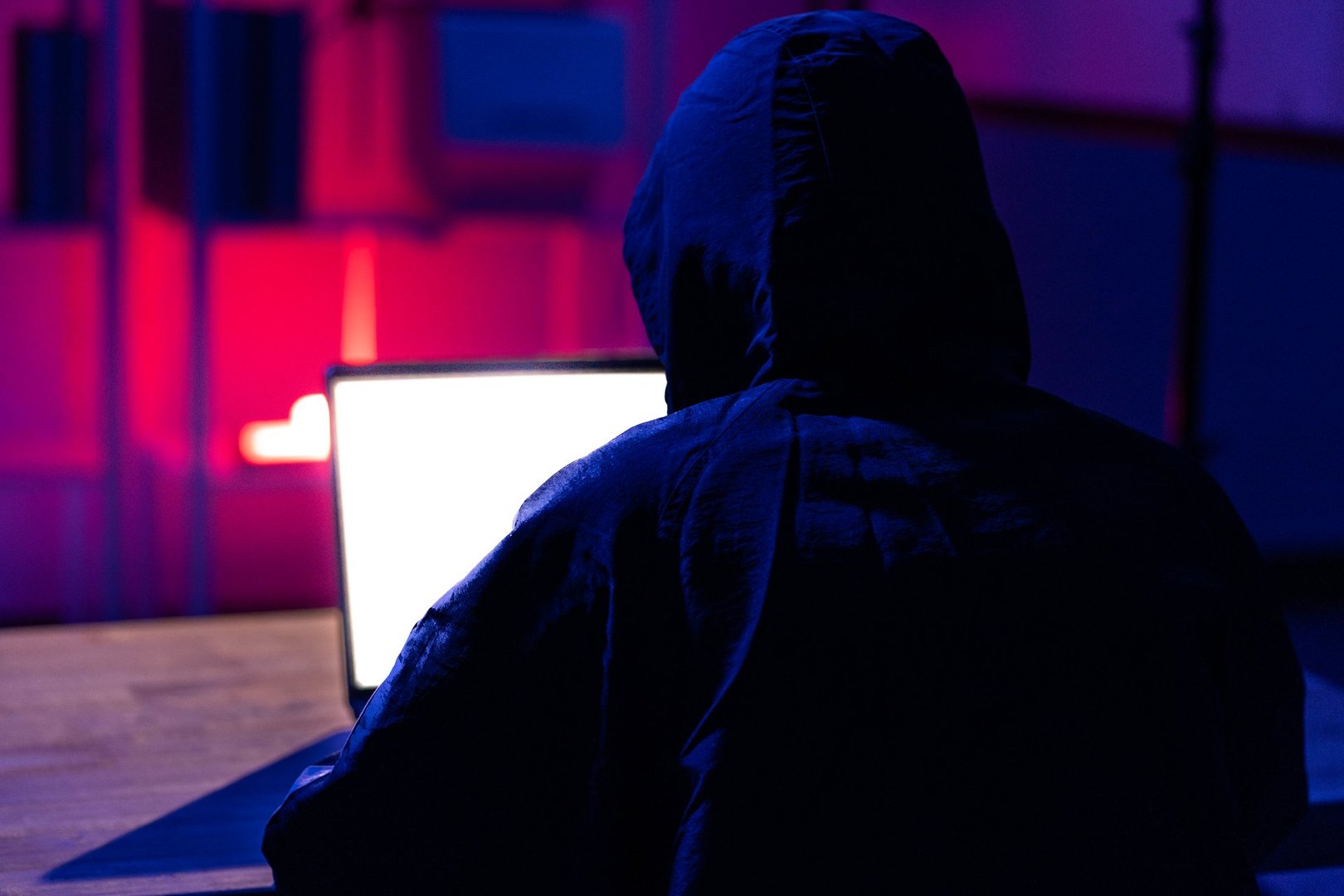 A young man in a black hoodie intently using a laptop in a neon-lit room, evoking themes of cybersecurity, hacking, coding, and digital privacy in a tech-driven world.