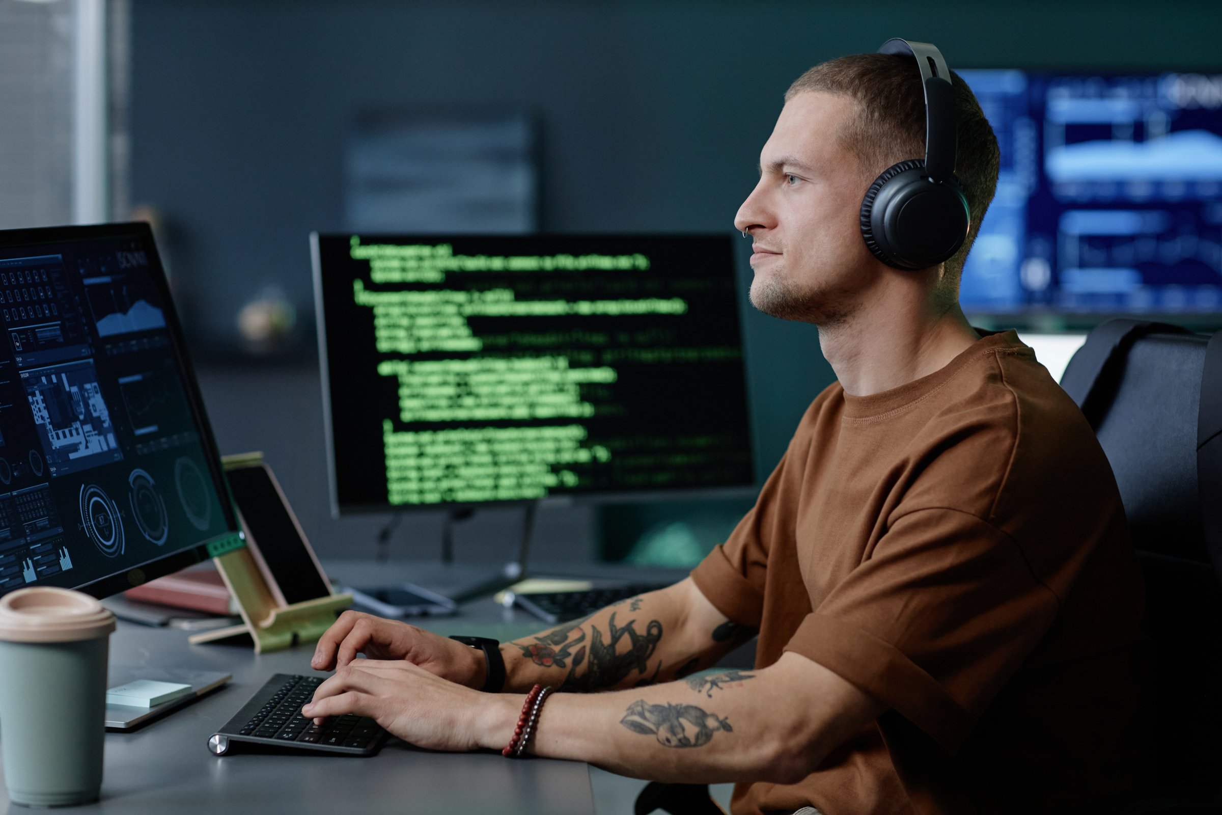 Side view of handsome male programmer wearing headphones working at ease on desktop computer writing code captured against multiple screens in corporate office