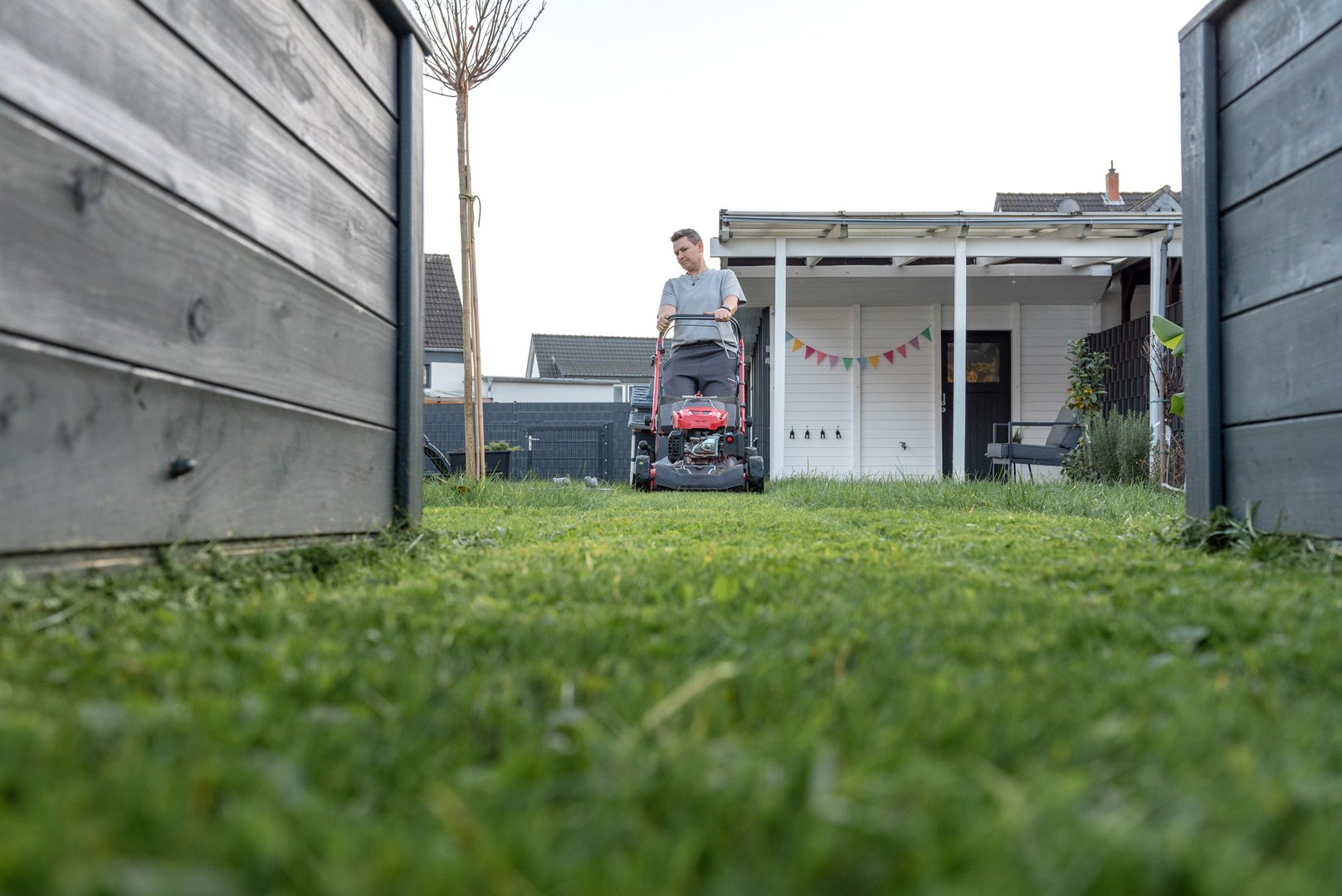 Man mowing grass in backyard with red lawnmower on a sunny day afternoon.