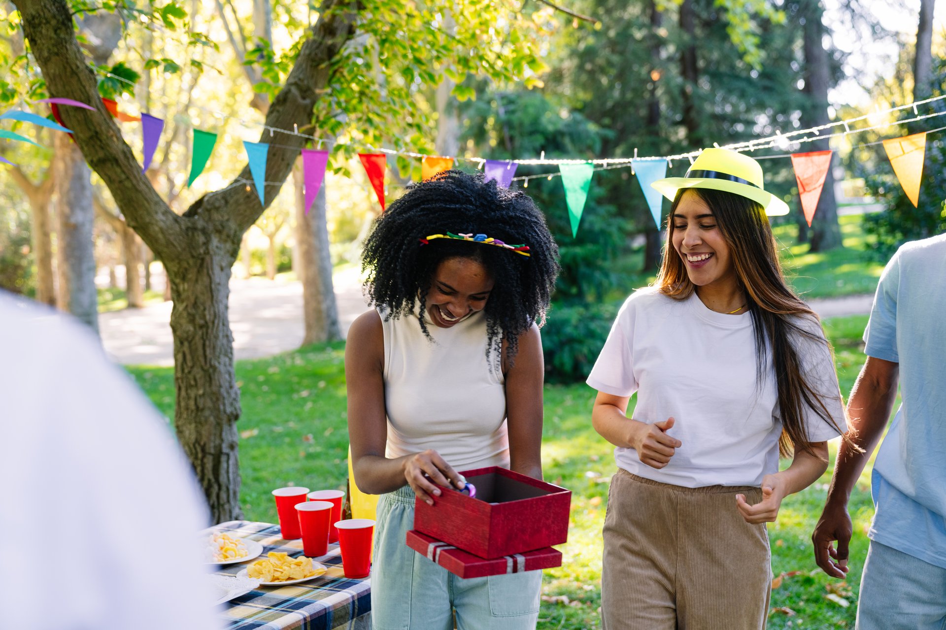 Young woman opening a gift surrounded by friends at her birthday party in a park, with colorful pennants hanging from the trees