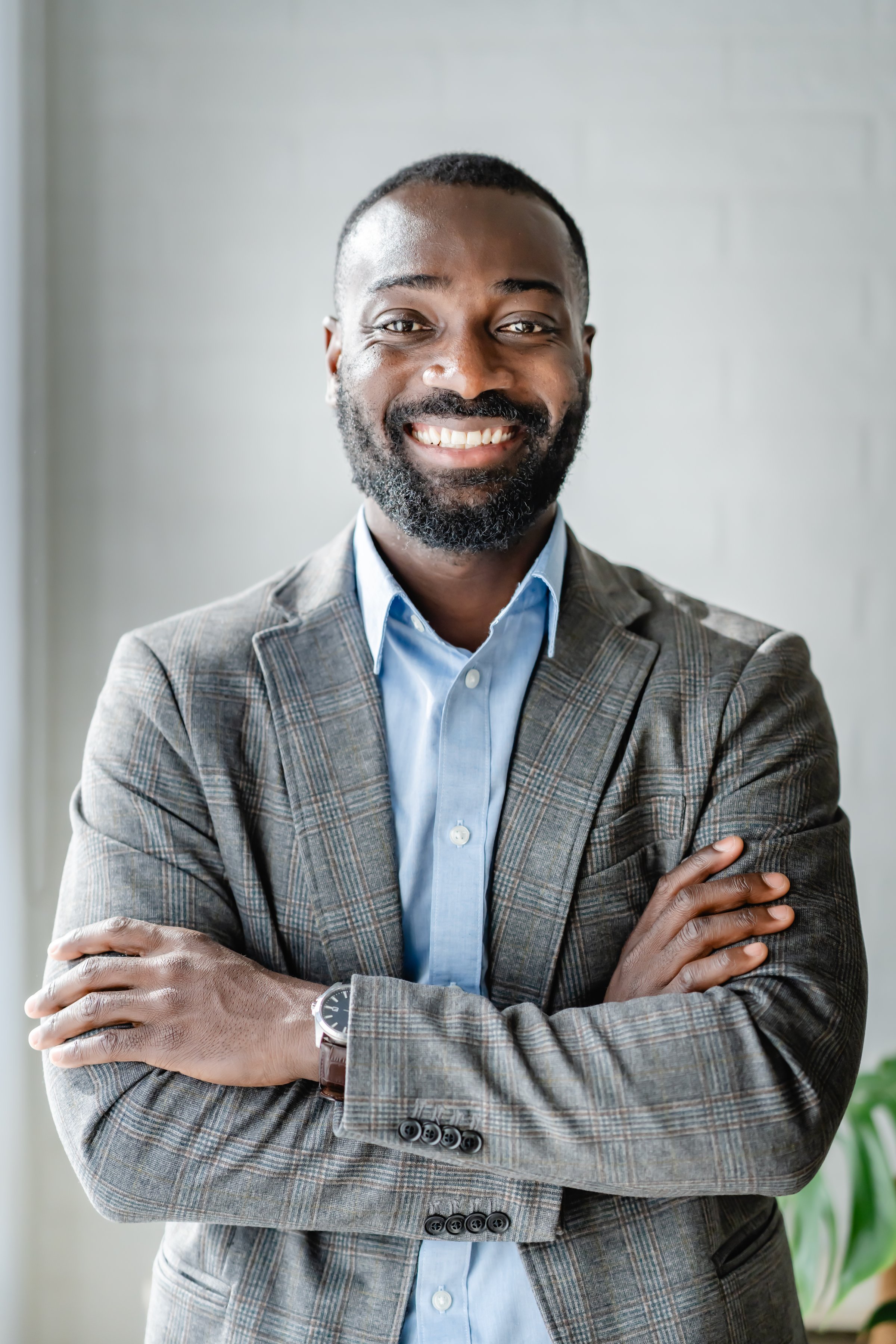 A confident African-American businessman stands with arms crossed, smiling at the camera. He is dressed in a gray blazer and blue shirt, exuding professionalism and warmth in a well-lit office space.