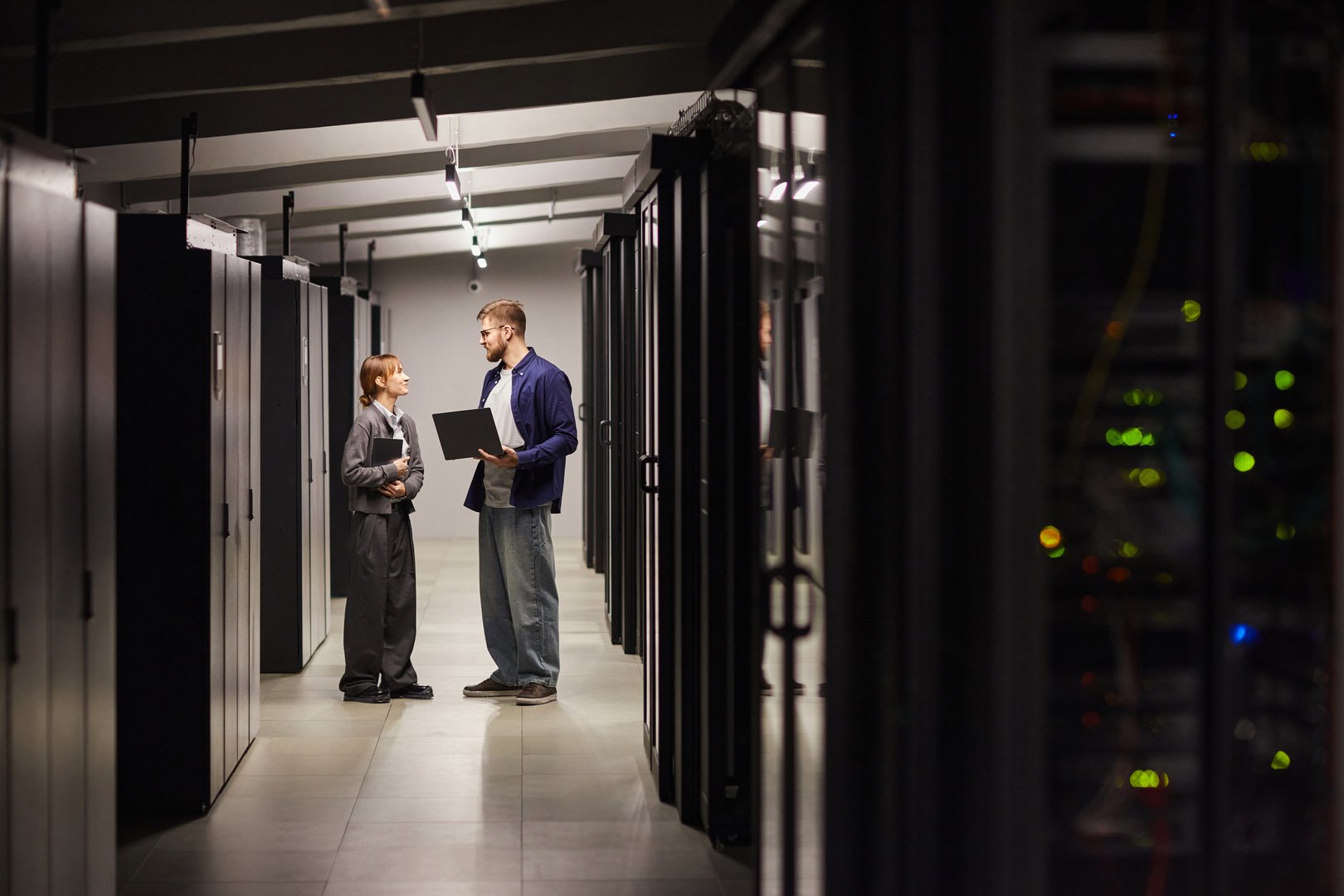 Two individuals discussing work in a well-lit, contemporary data center while holding laptop. Environment consists of multiple server racks and advanced technology