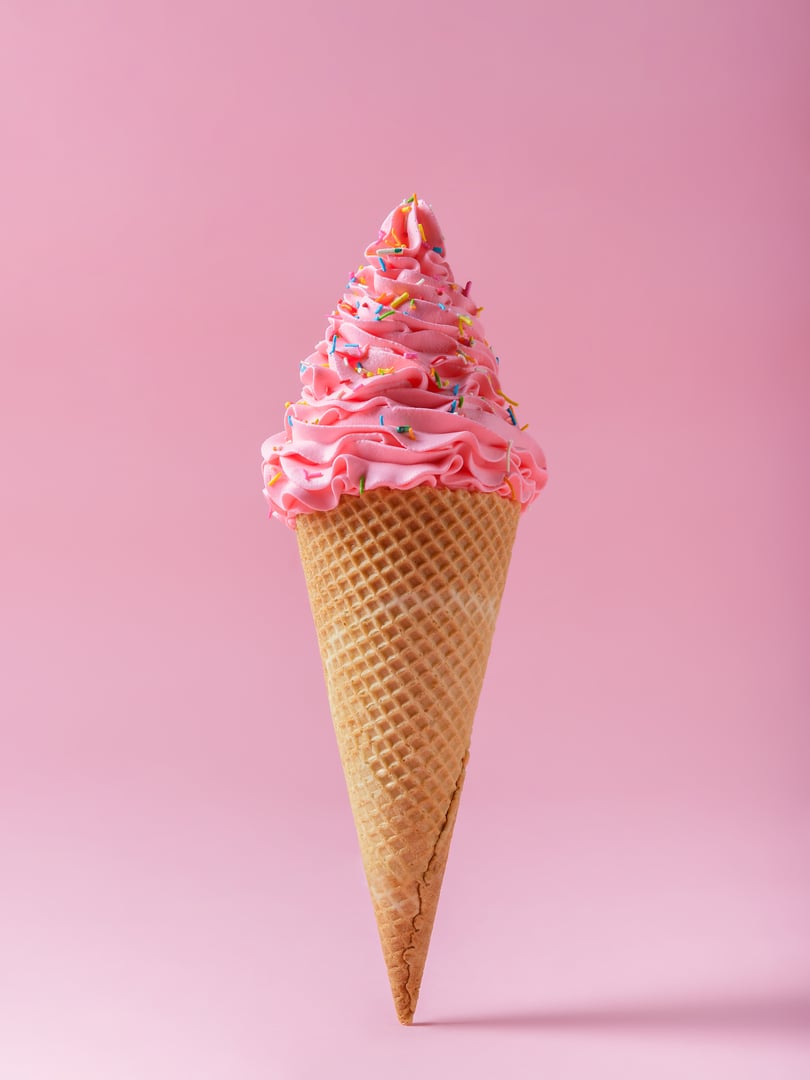 A tall ice cream cone filled with fluffy pink ice cream and colorful sprinkles stands against a pale pink background.