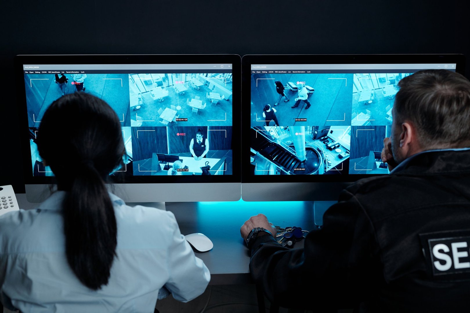 Rear view of male and female security guards sitting in front of desktop computers with video camera on screens in surveillance room