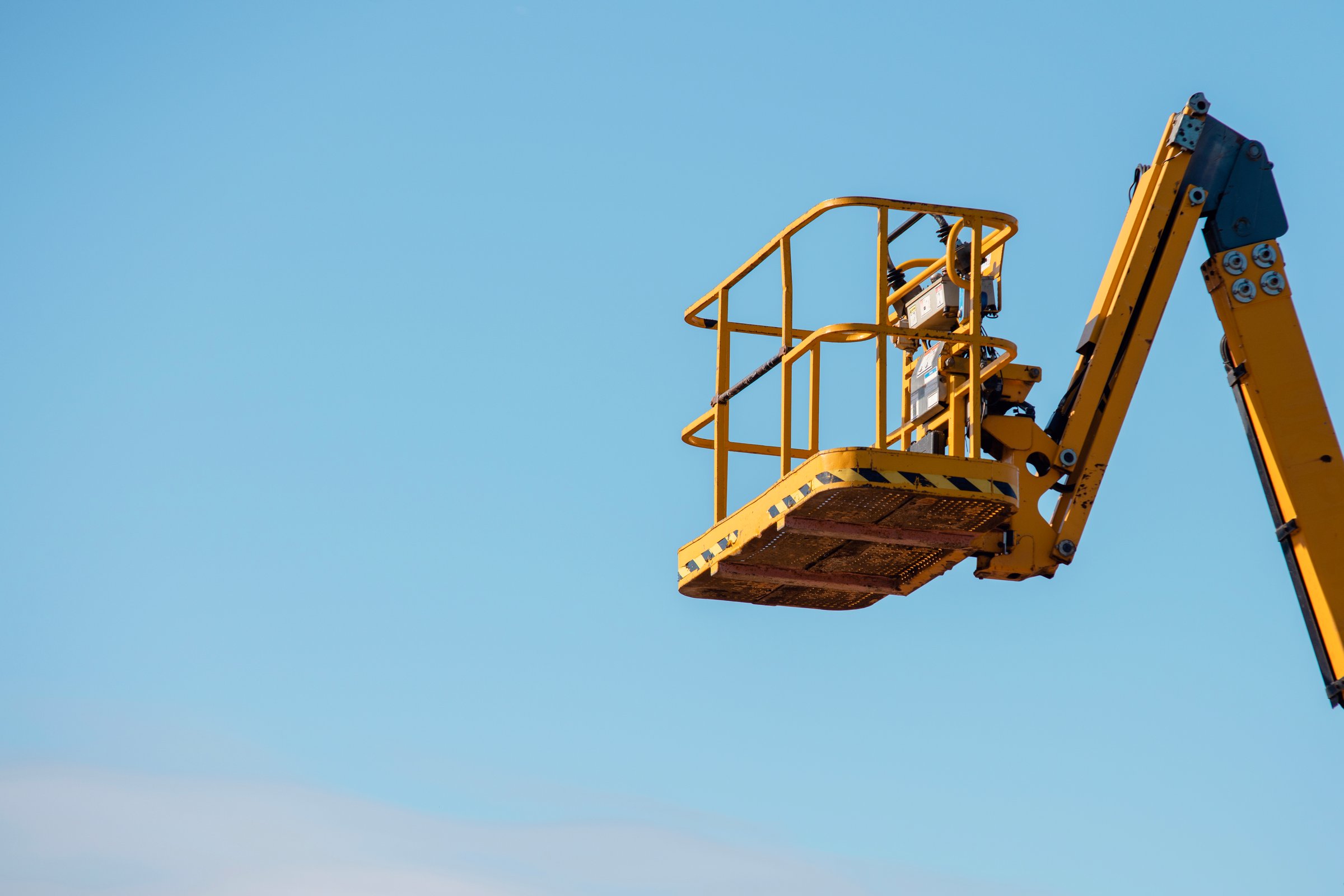 Elevated boom lift platform cherry picker against clear blue sky during daytime works on construction site with bright sunlight illuminating the machinery