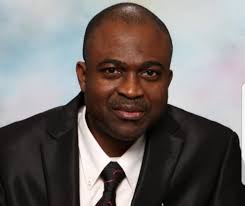 A person wearing a black suit and tie, set against a neutral background, with a calm expression.