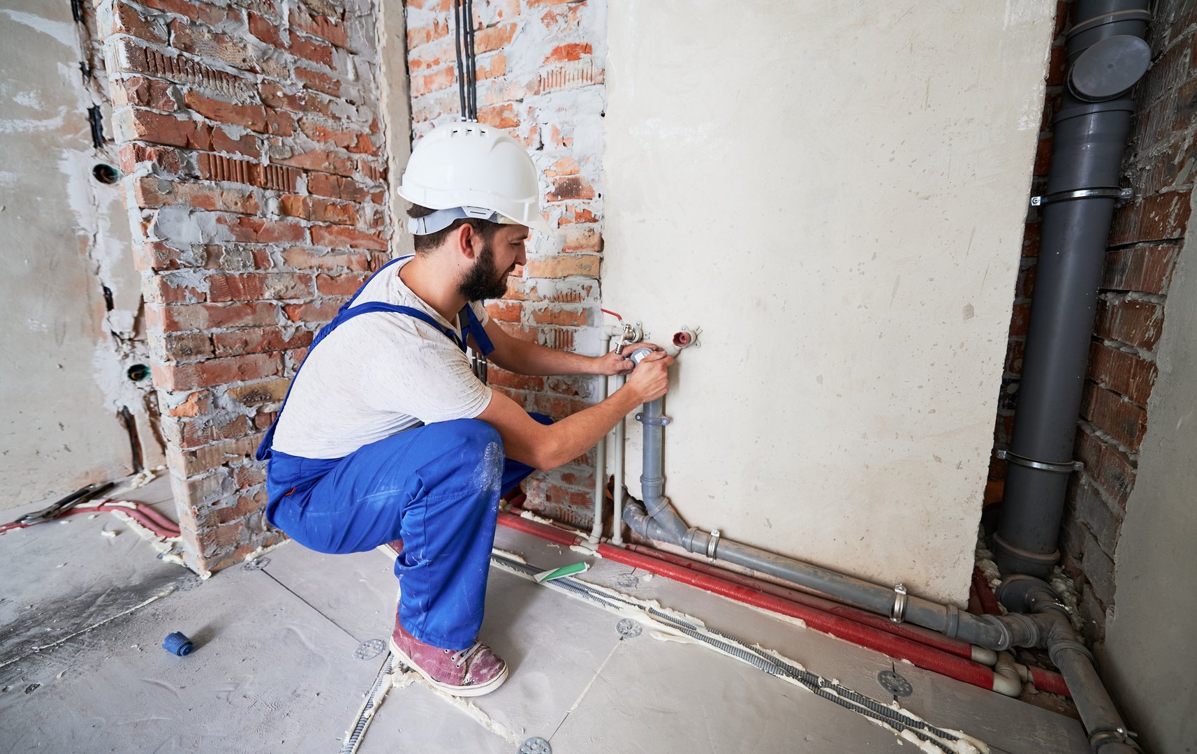 Bearded young man in work overalls installing heating pipes in apartment. Male worker in safety helmet doing maintenance jobs for water and heating systems. Concept of plumbing and home renovation.