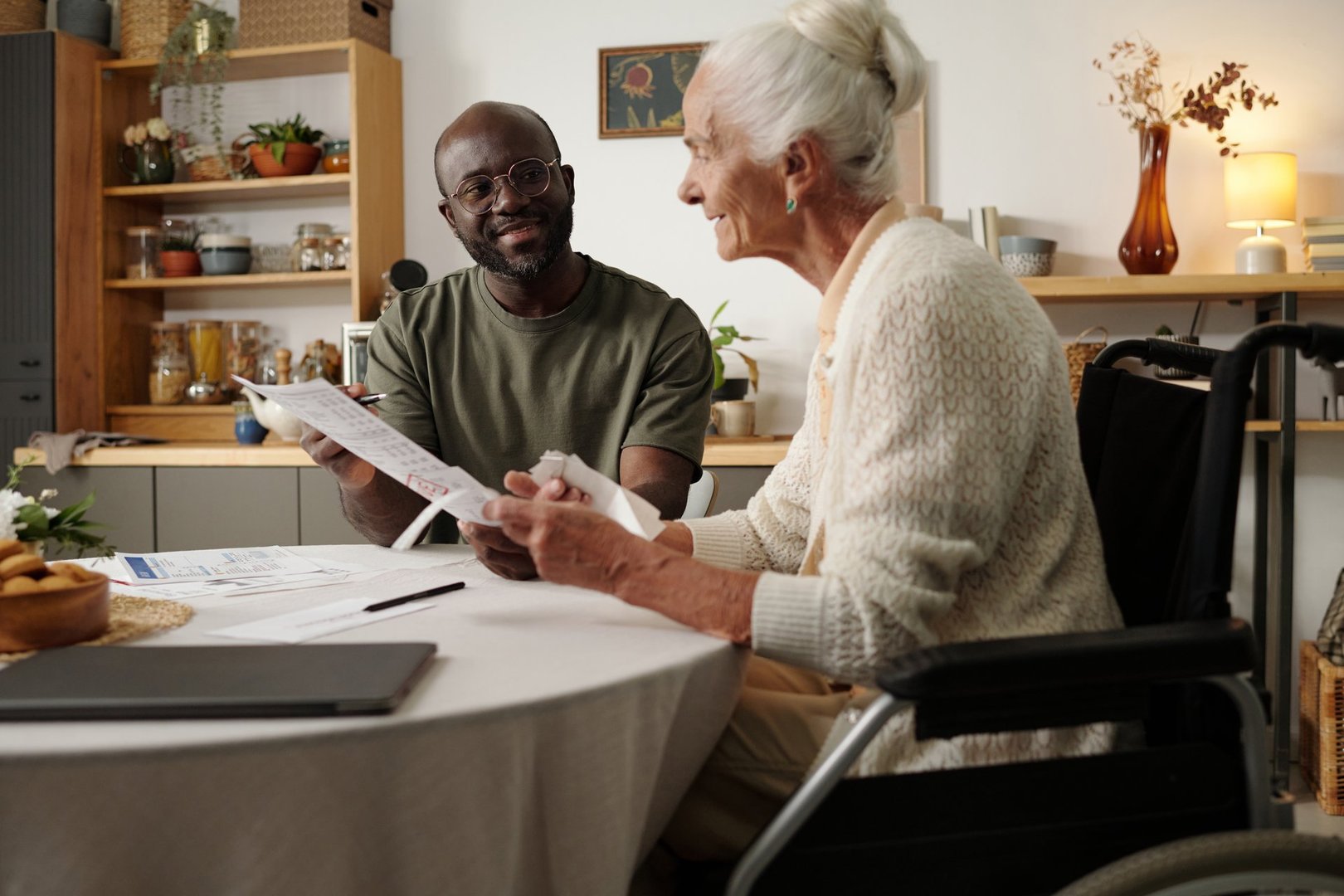 Black man explaining documents to senior Caucasian woman with disability in wheelchair, both sitting at table in home setting, engaging in conversation and paperwork
