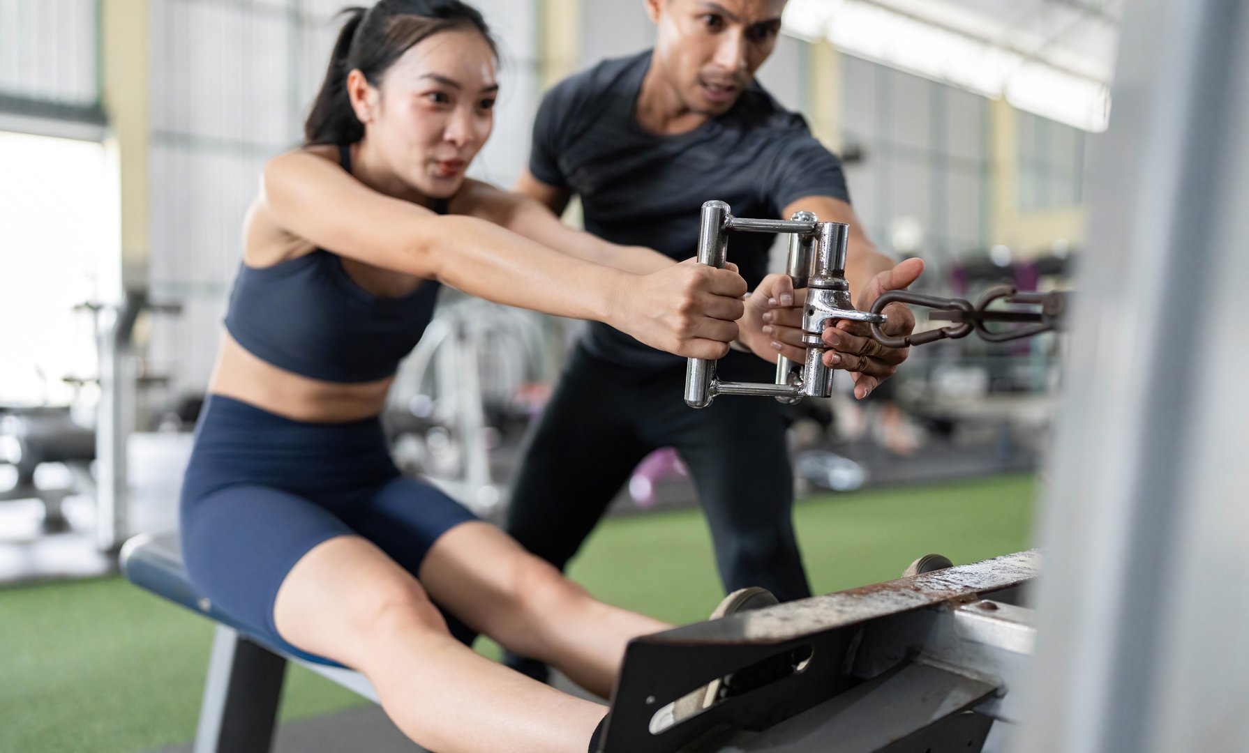 A personal trainer helps a female client during a rowing machine workout, focusing on form and motivation in a gym environment.