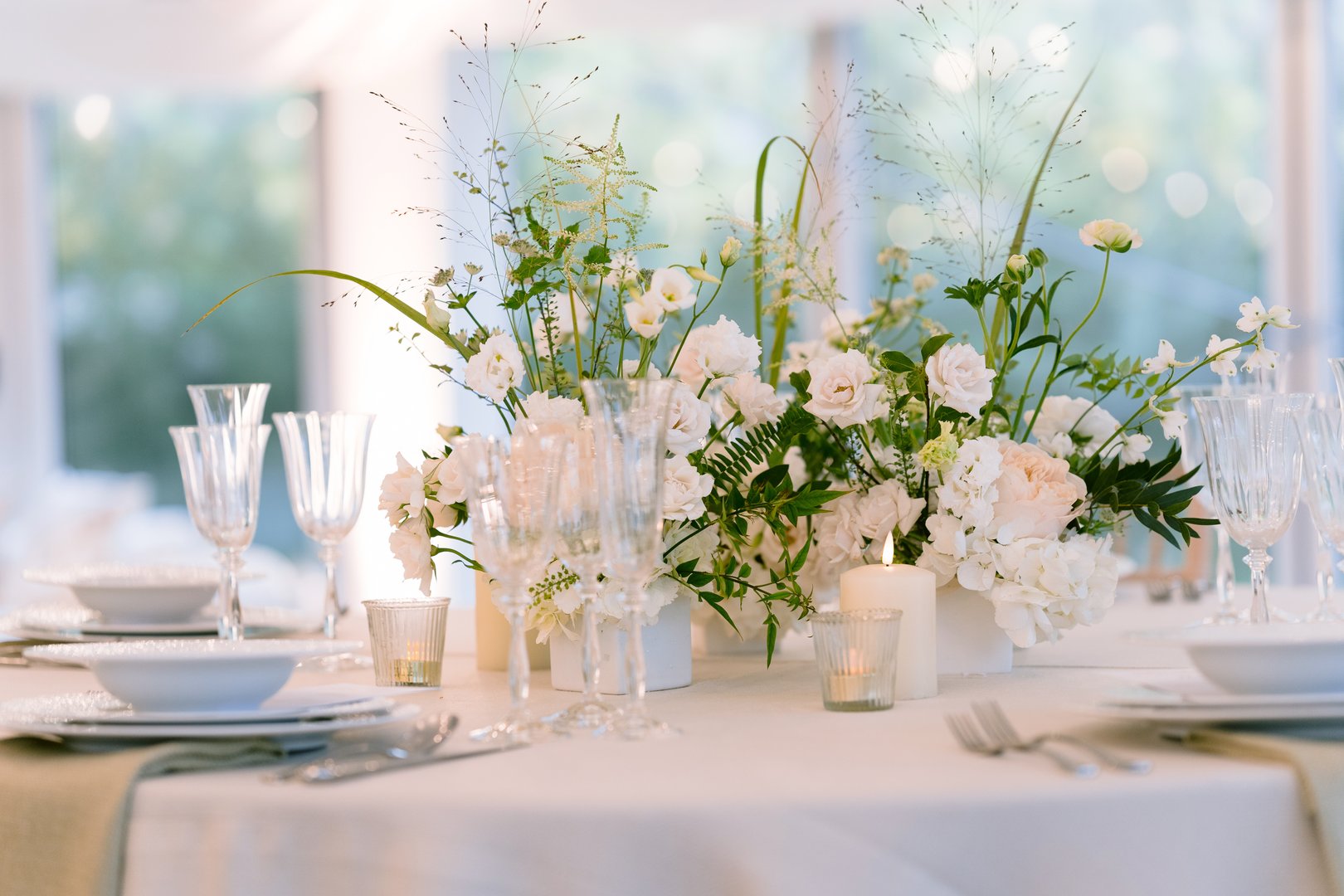 Elegant floral decoration on the back of a light upholstered chair at an indoor wedding ceremony. The arrangement features soft blush and ivory roses, subtle greenery, and a trailing silk ribbon, creating a romantic and sophisticated detail. Set within a refined indoor venue with warm lighting and a clean, minimalist aesthetic, the scene exudes timeless charm—perfect for modern, classic, or intimate wedding inspiration