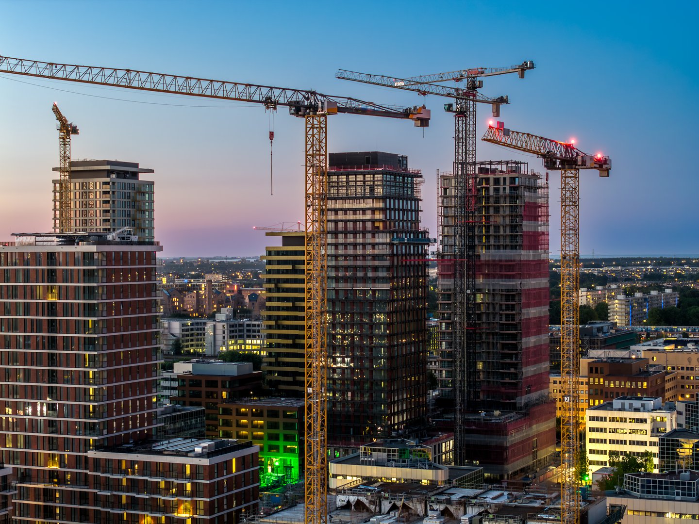 Evening aerial view of the Zuidas business district in Amsterdam South, where multiple high-rise towers are under construction. Tower cranes rise above illuminated glass facades as city lights shimmer against a fading sky, highlighting rapid urban growth, real-estate investment, and the Netherlands' modern architectural ambitions.