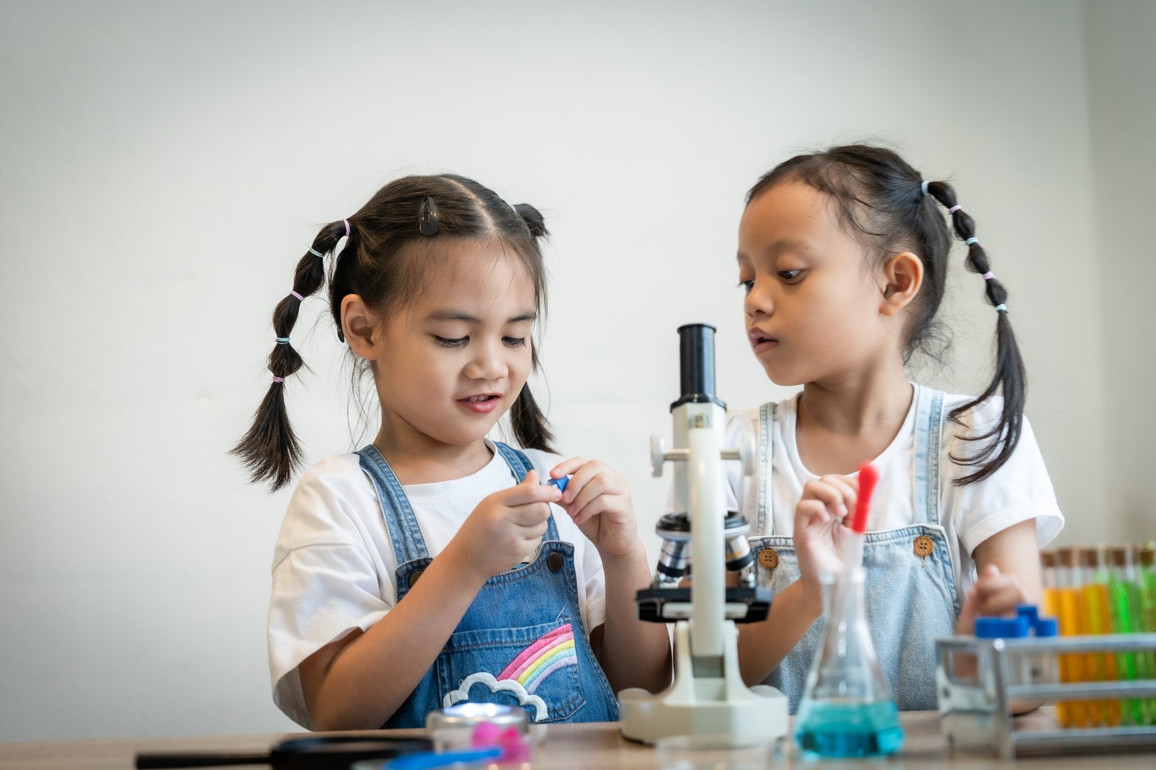 Two young girls are playing with a microscope, looking at a slide. The slide has a small object on it, and the girls are examining it closely. The scene is playful and educational