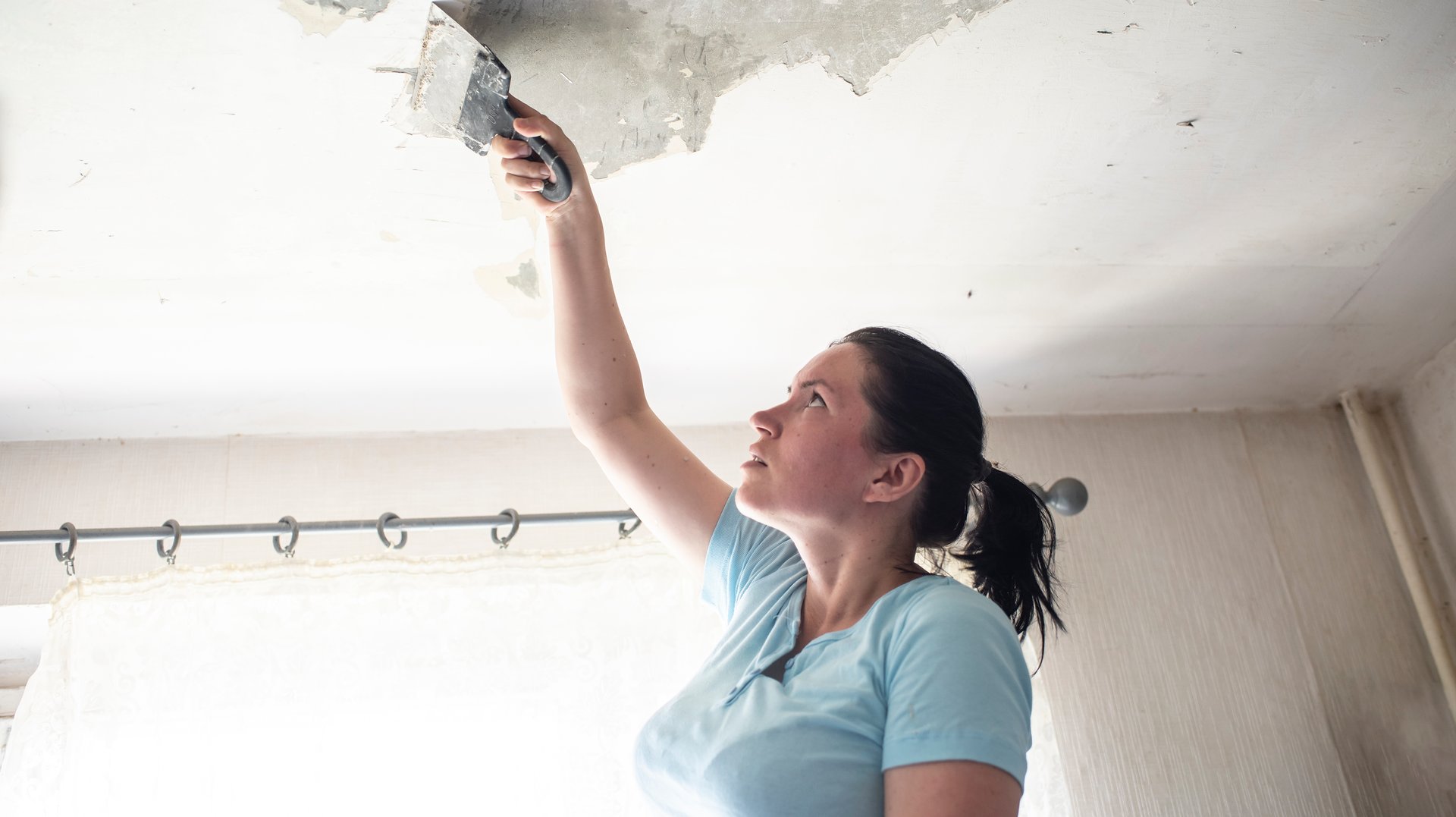 a woman using a spatula removes the top layer of old paint on the ceiling